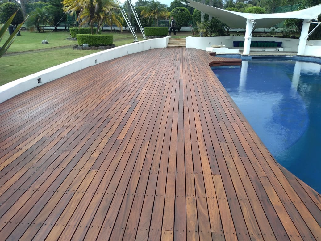 Un paseo marítimo de madera junto a una piscina azul en un parque ajardinado, con un toldo blanco encima.