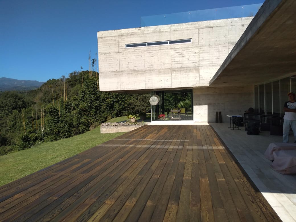 Casa moderna con terraza de madera y patio cubierto con vistas a una ladera verde bajo un cielo azul.
