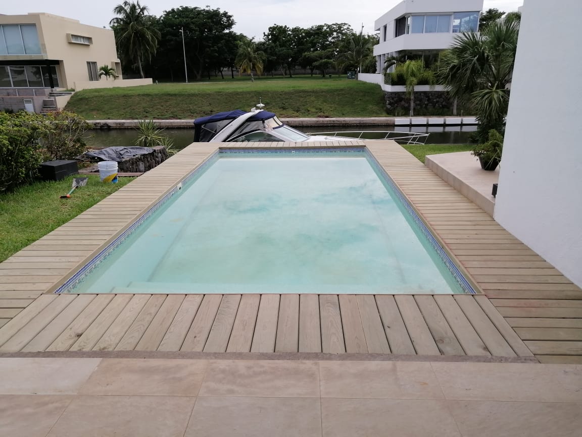 Piscina rectangular en el patio trasero con agua azul pálida, terraza de madera y casas al fondo.