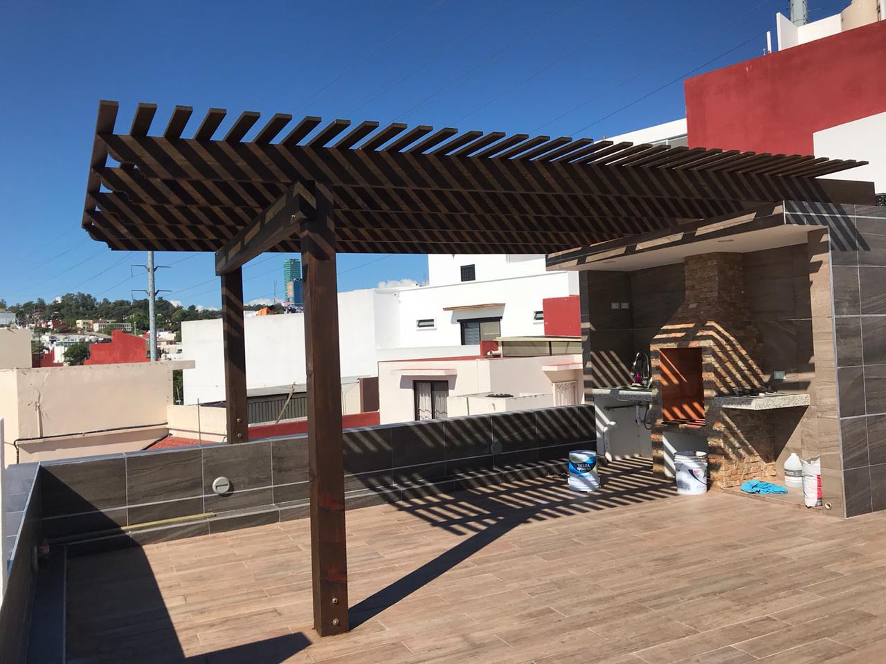 Pérgola de madera exterior con techo de listones que proyecta sombras en una terraza en la azotea bajo un cielo azul.