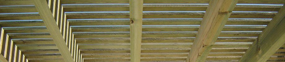 Las vigas de madera de una pérgola proyectan sombras rayadas sobre un cielo azul y las copas verdes de los árboles.