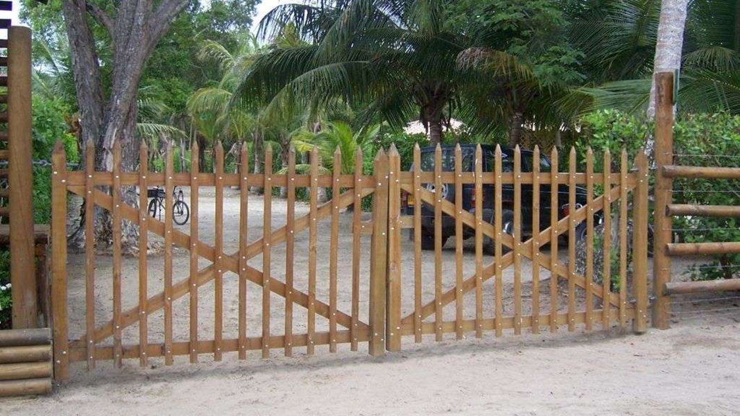 Una puerta de listones de madera cruza un camino de arena, rodeado de árboles y vegetación.