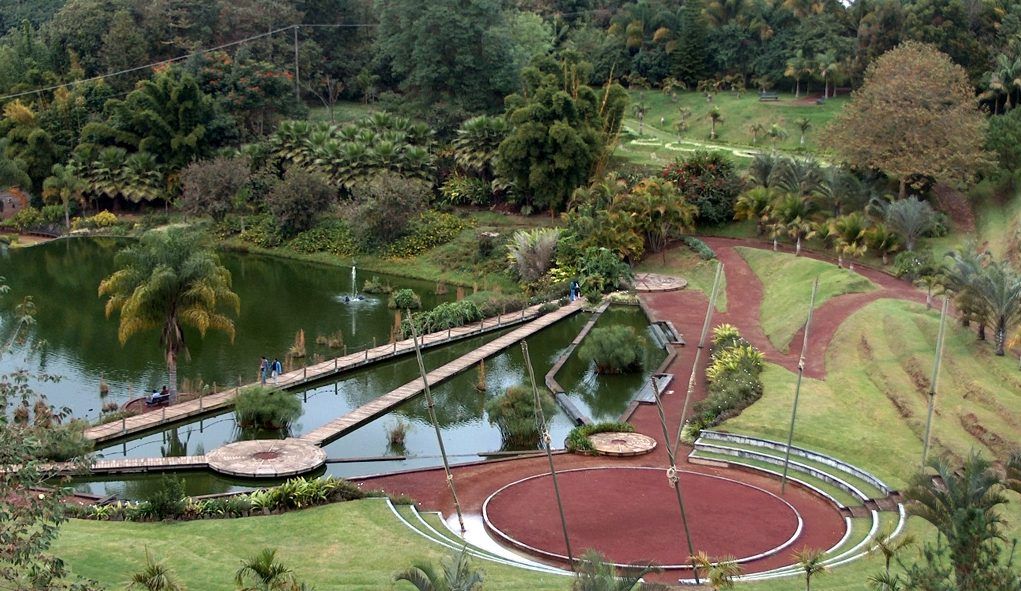 Parque tropical con estanque, puente, sendero circular rojo y exuberantes jardines en la ladera.