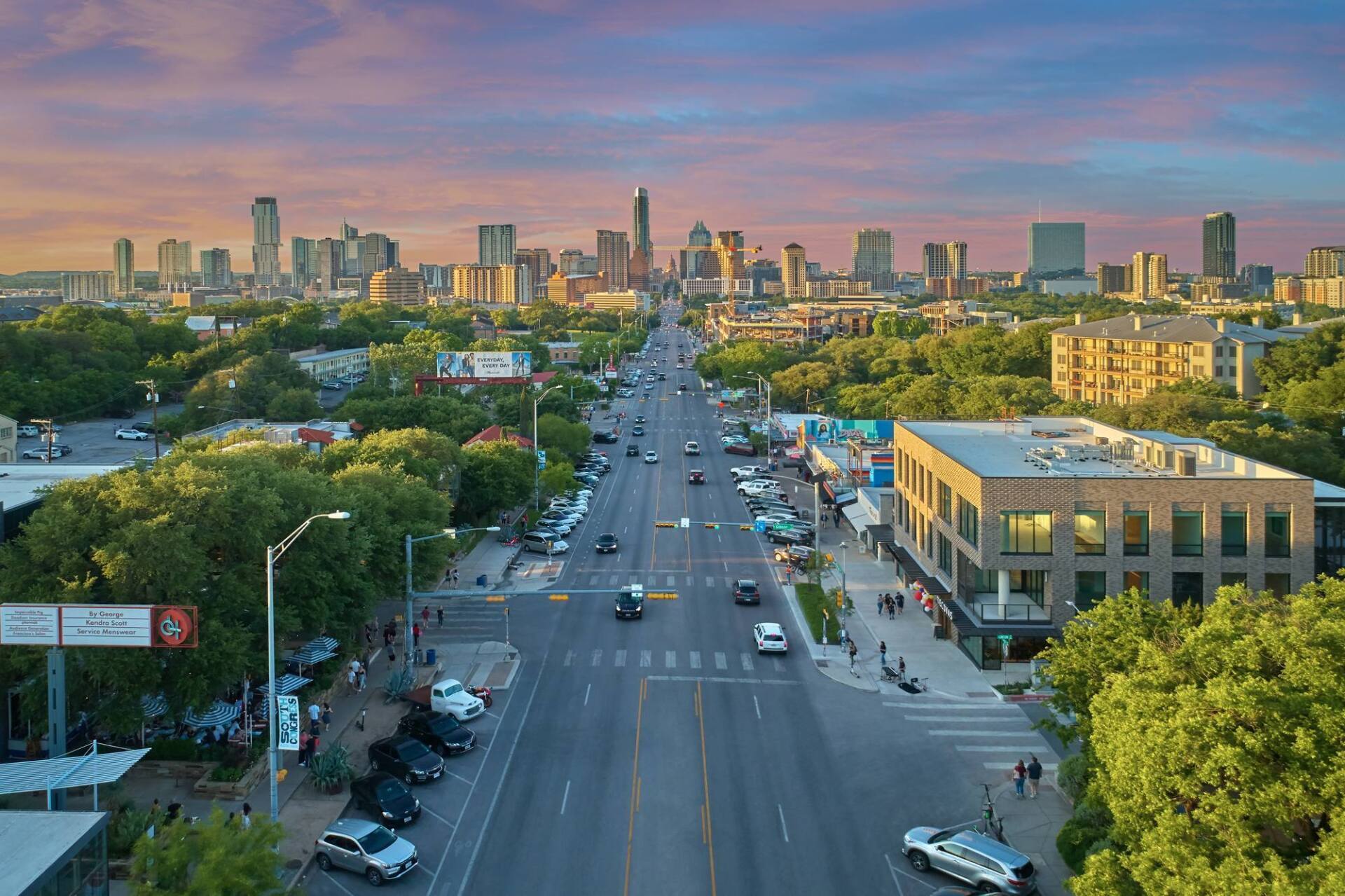 City street with buildings, cars, and trees, with a skyline at sunset.