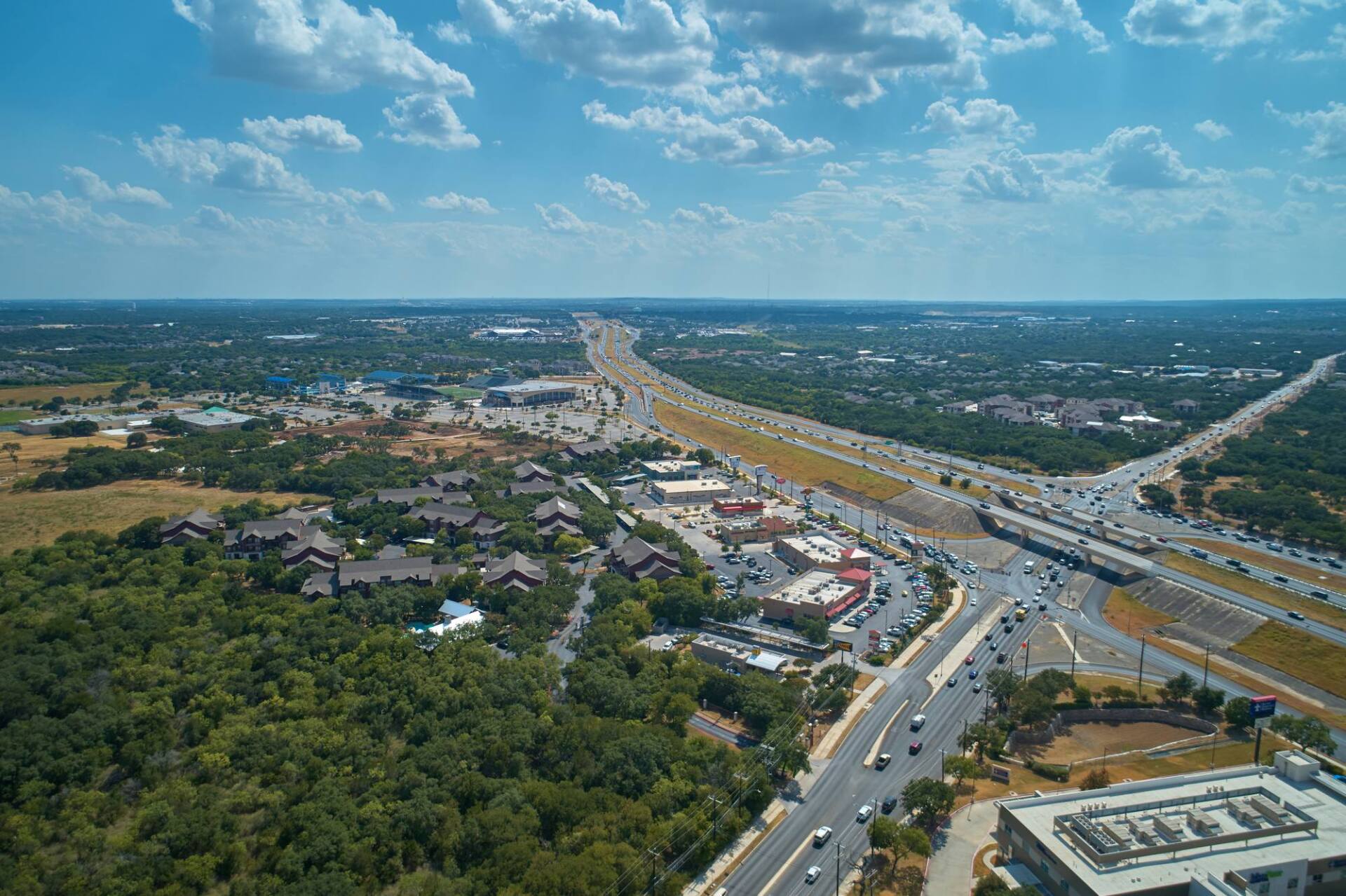 Aerial view of a city with highways, buildings, and green trees under a blue sky with clouds.