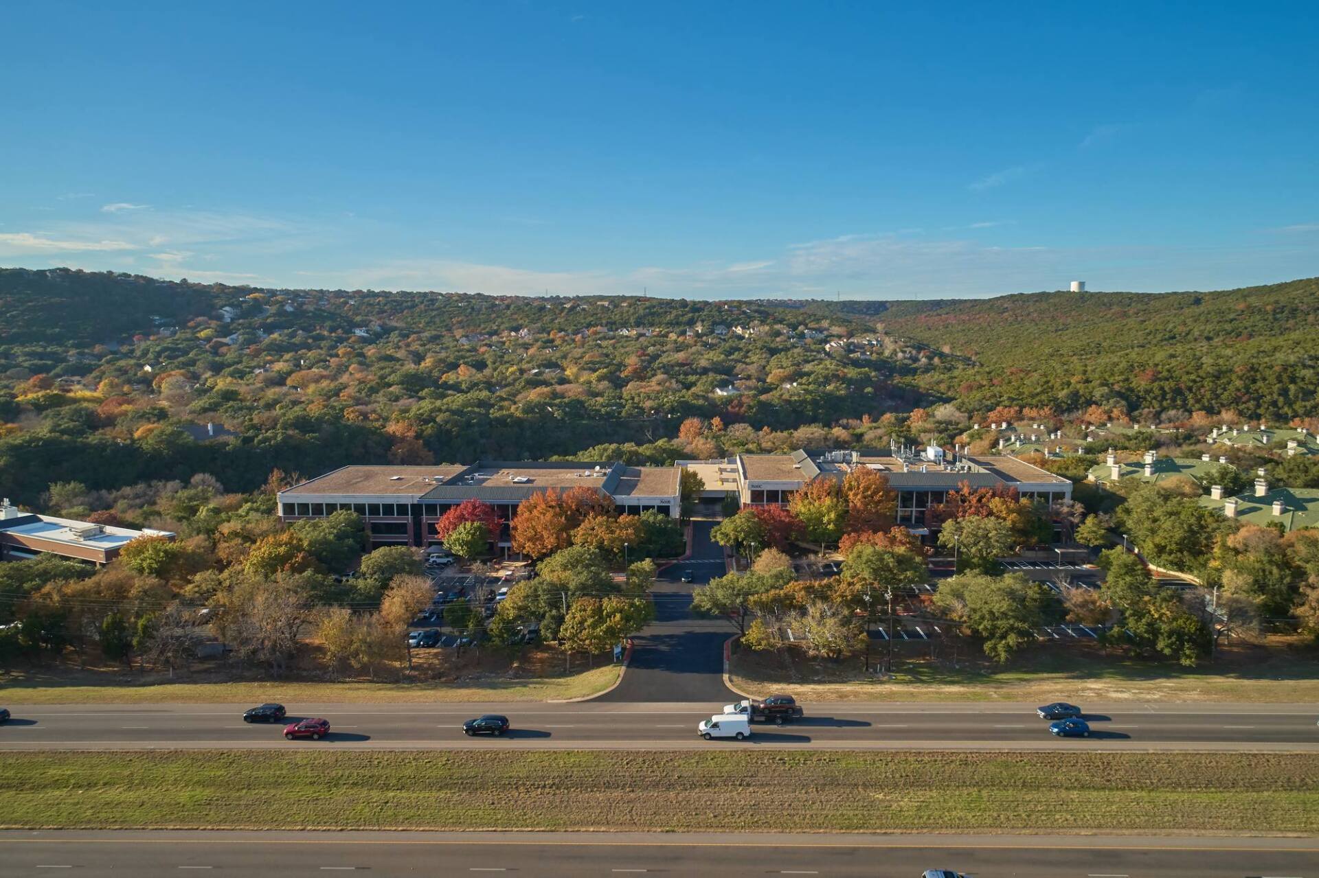 Office building surrounded by fall foliage, highway in foreground, blue sky.