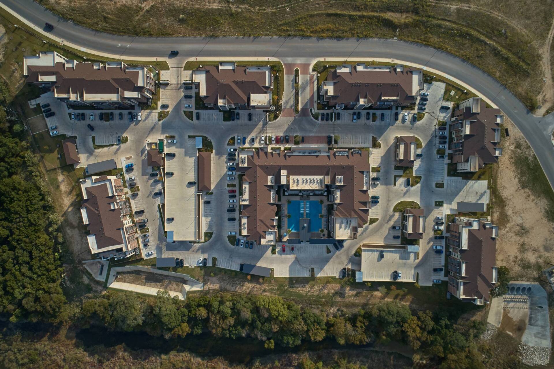 Aerial view of apartment complex with a pool, parking, and surrounding road and green space.