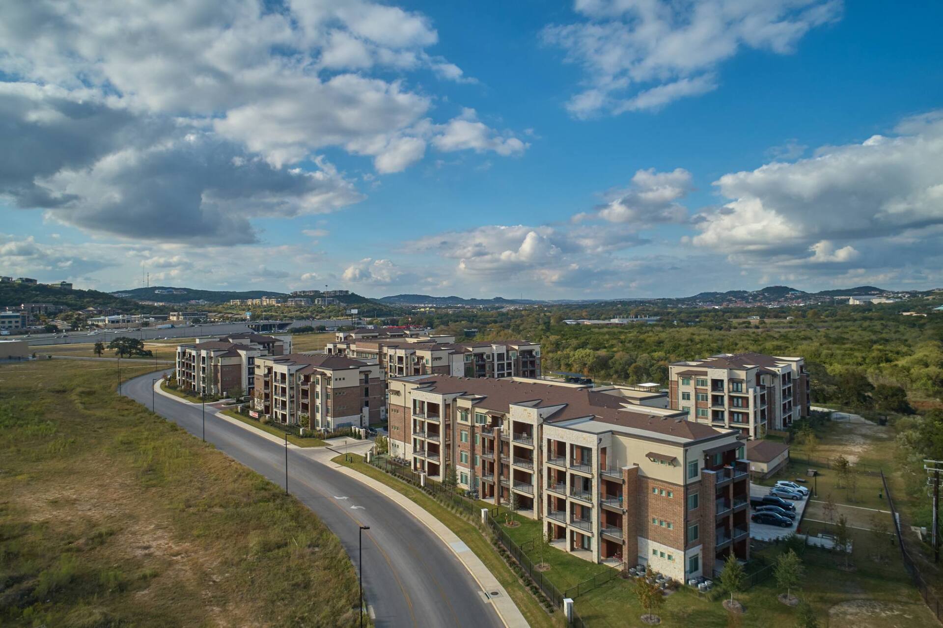 Apartment buildings with brown brick accents, set against a green landscape under a cloudy blue sky.
