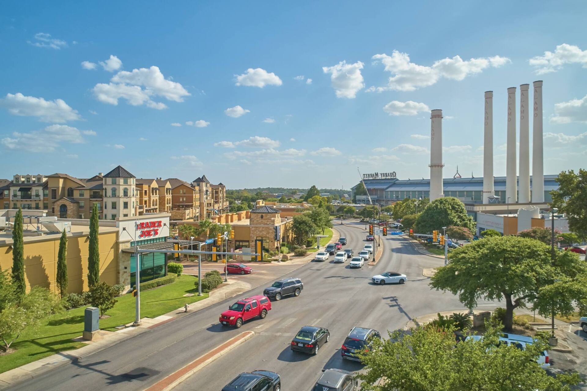 Road with cars, shops, and buildings under a blue sky with white clouds. Power plant smokestacks are in the distance.