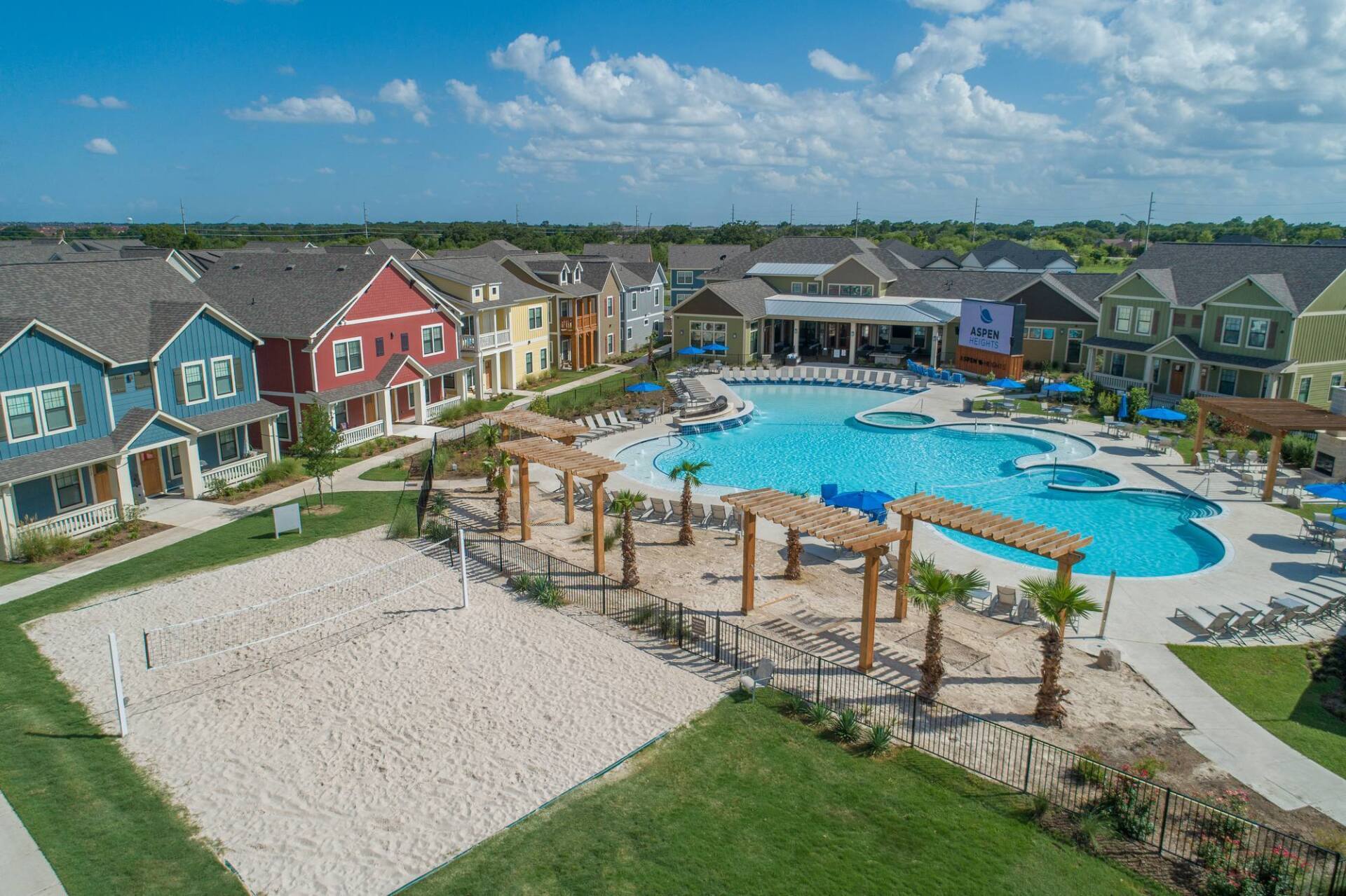 Aerial view of a colorful residential complex with a large pool, volleyball court, and blue sky.