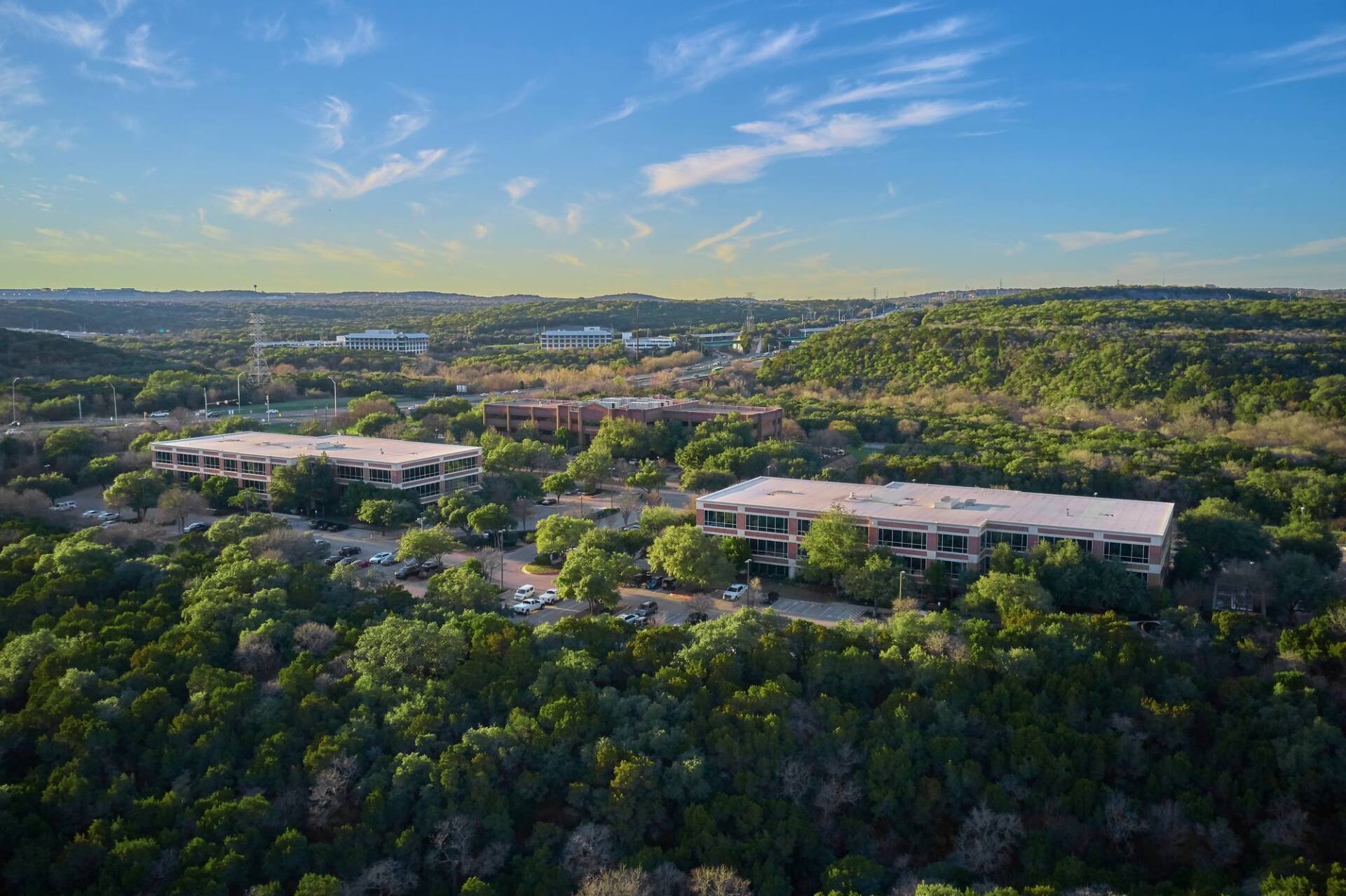 Aerial view of office buildings amidst lush green trees under a blue sky with clouds.