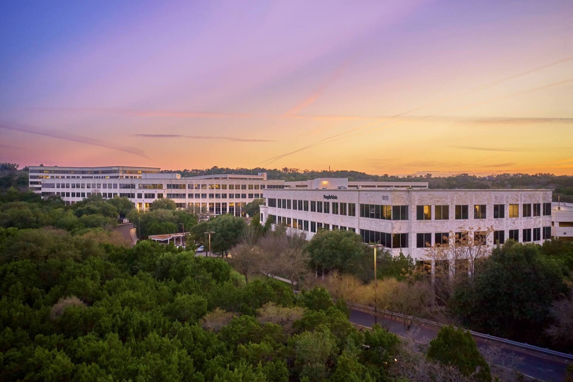 Office buildings at dusk, viewed from above a tree line, with a colorful sky.