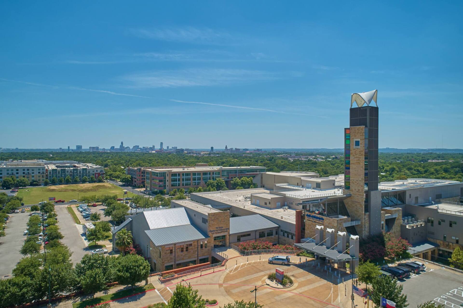 Modern buildings and tower against a skyline, under a blue sky, surrounded by trees and a parking lot.