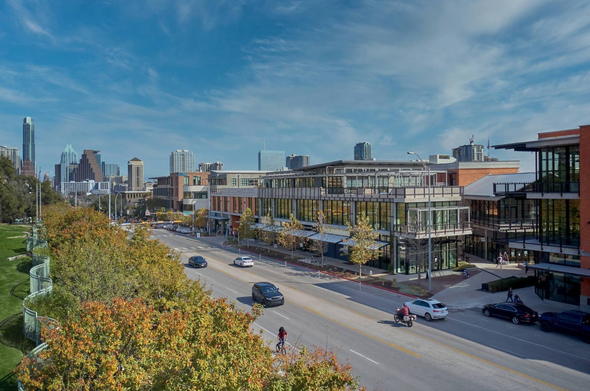 Street view of buildings, road with cars, and trees against a city skyline under a blue sky.