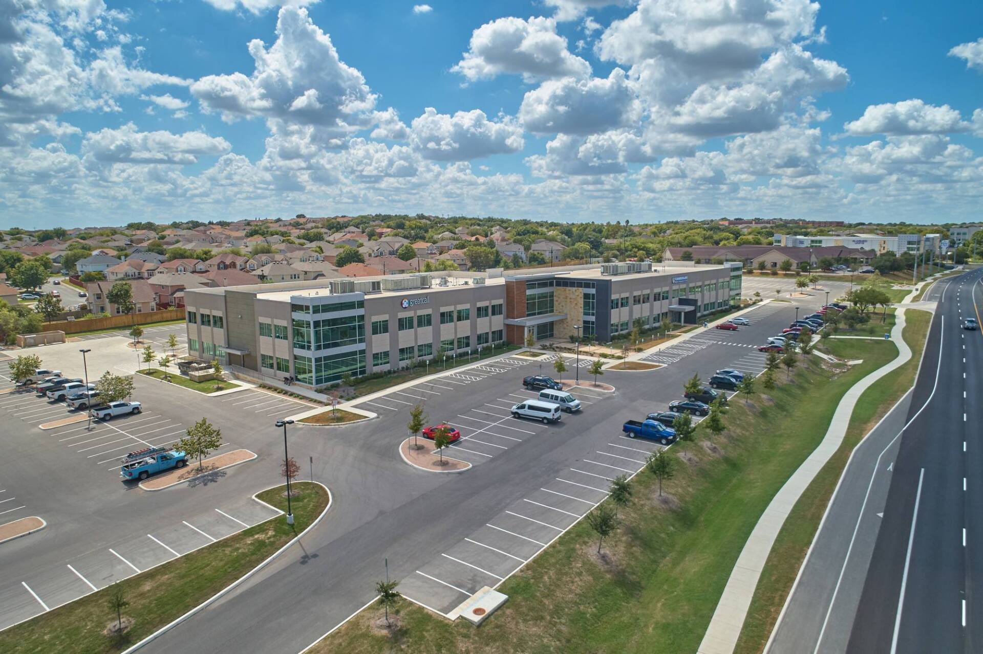 Aerial view of a modern office building with parking, a highway, and residential homes under a blue sky.