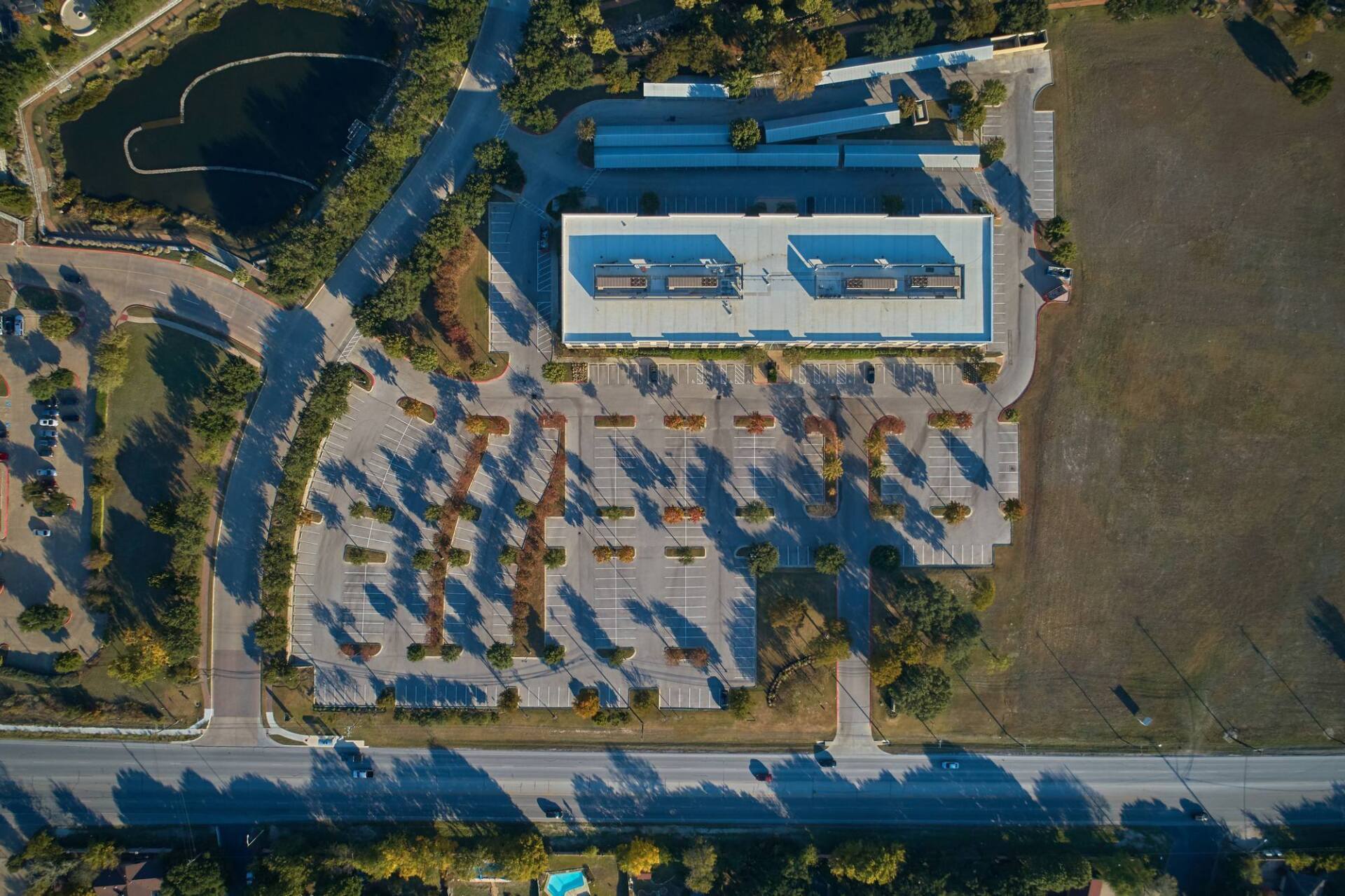 Aerial view of a building with a parking lot and a pond. Trees cast long shadows on the lot.