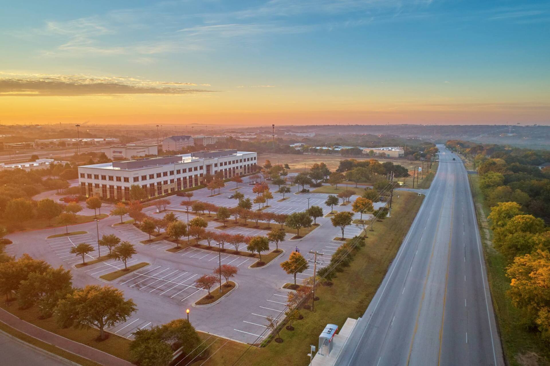Aerial view of a building with a large parking lot, and a long road at sunrise with a blue and orange sky.