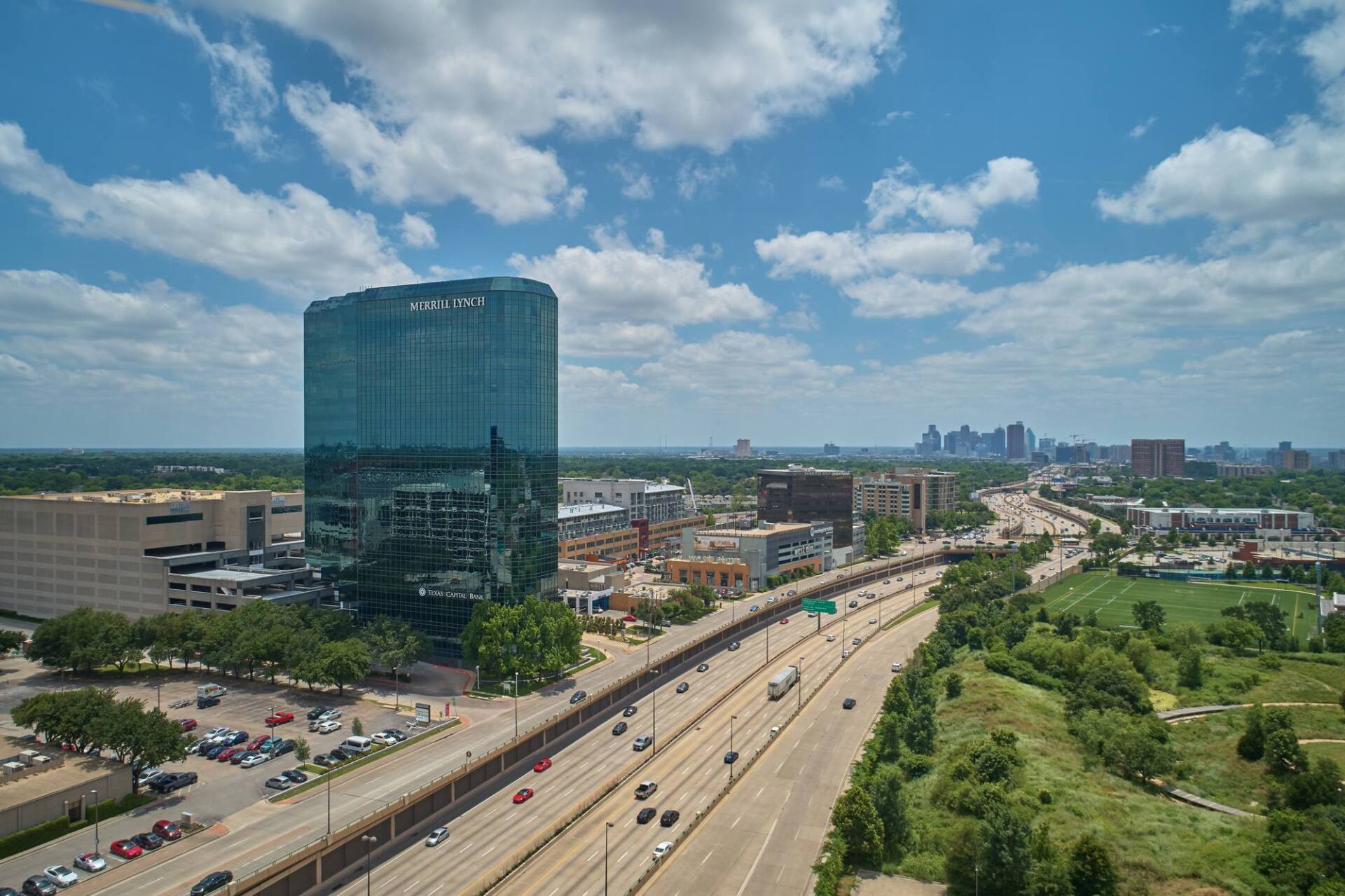 Blue glass skyscraper, highway, and city skyline under a partly cloudy sky.
