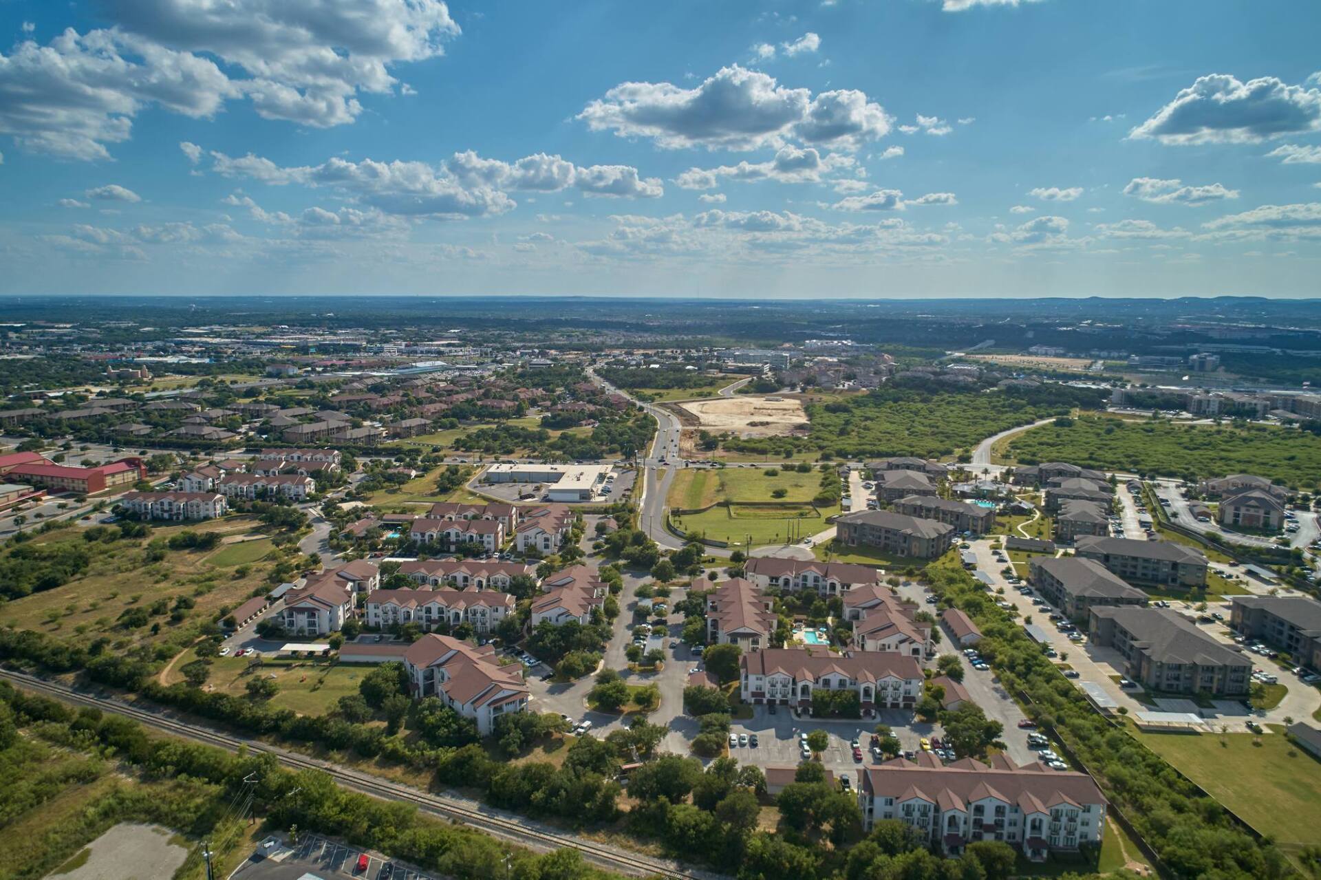 Aerial view of a suburban neighborhood with buildings, trees, a road, and blue sky with clouds.