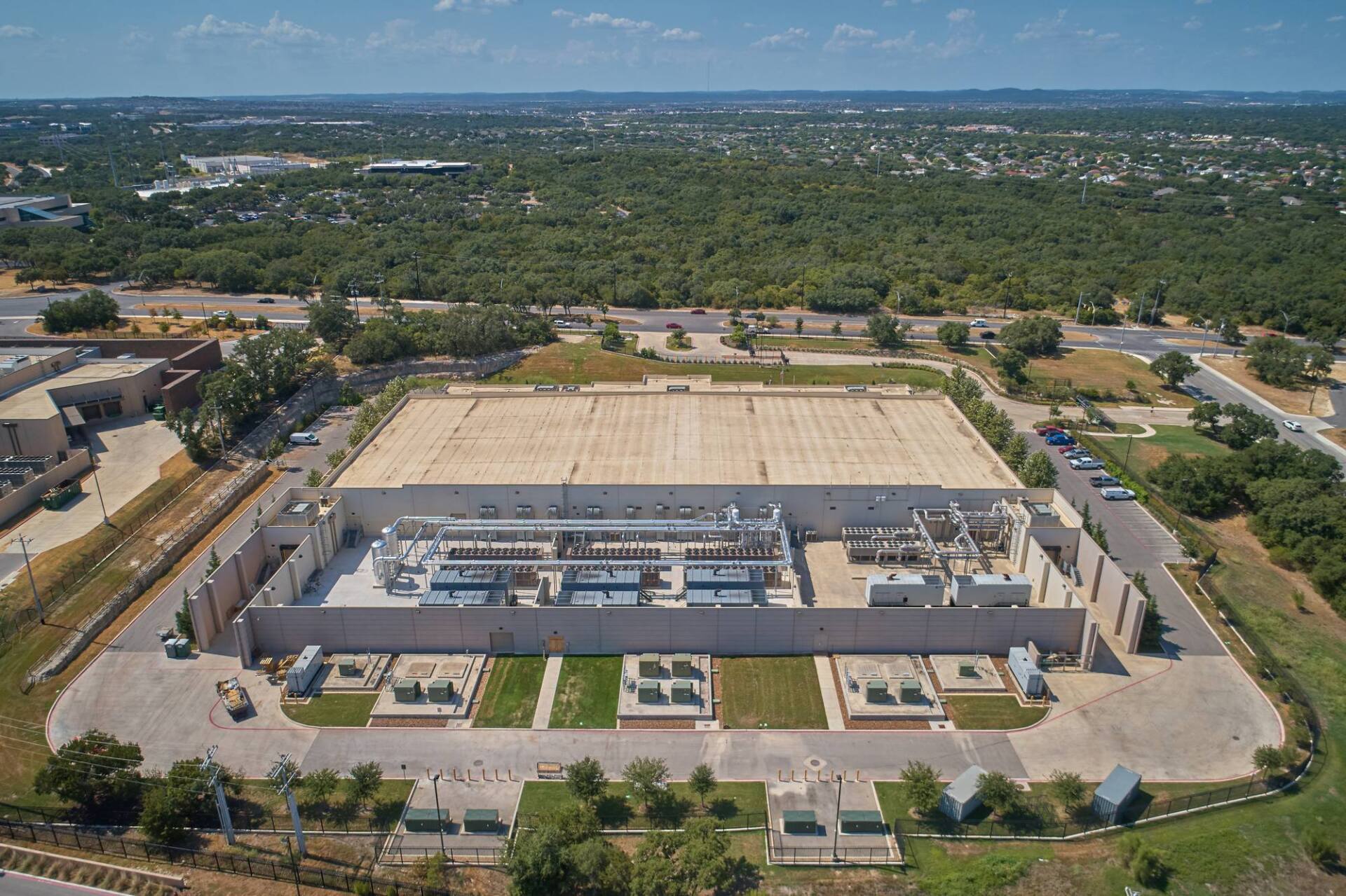 Aerial view of a large, flat-roofed data center in an urban area, surrounded by vegetation and roads.