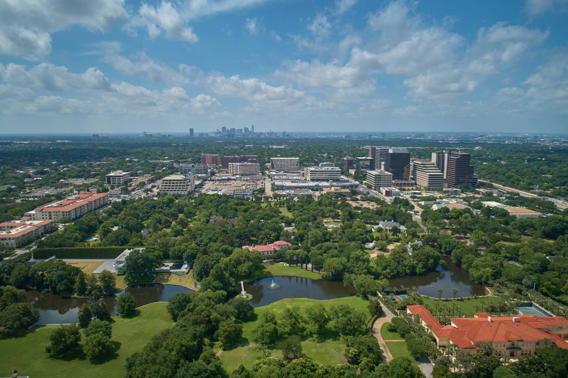 Aerial view of a city with a park, buildings, and a distant skyline under a blue sky.