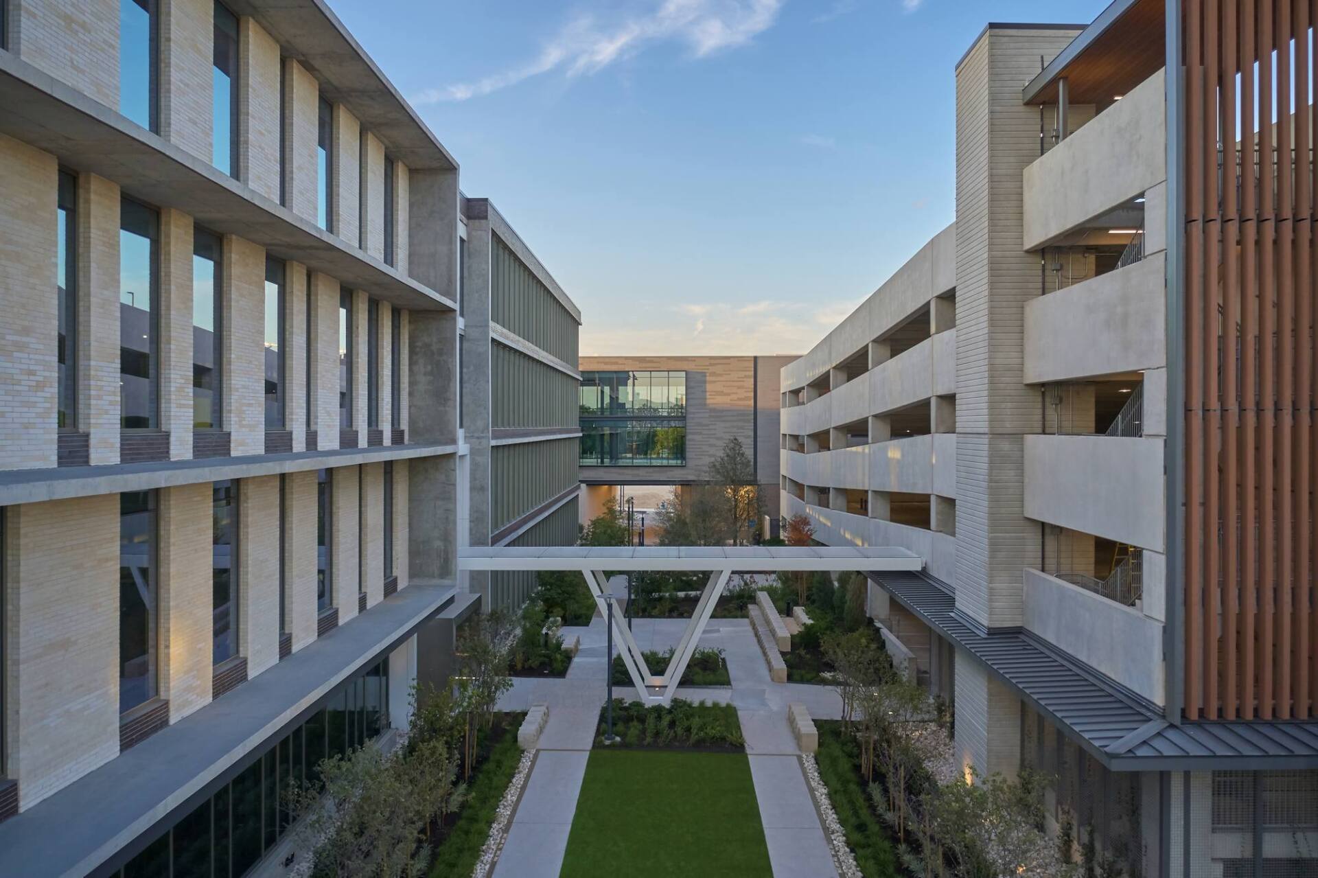 Modern buildings connected by walkways over a green courtyard with trees. Sky visible.