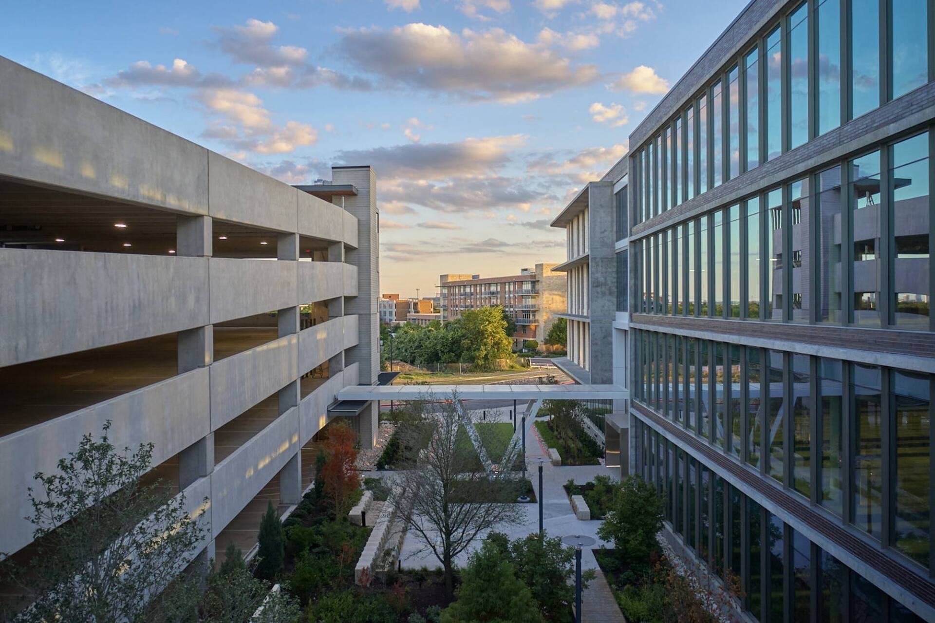 Two modern buildings flank a courtyard with trees, skybridge, and parking garage.
