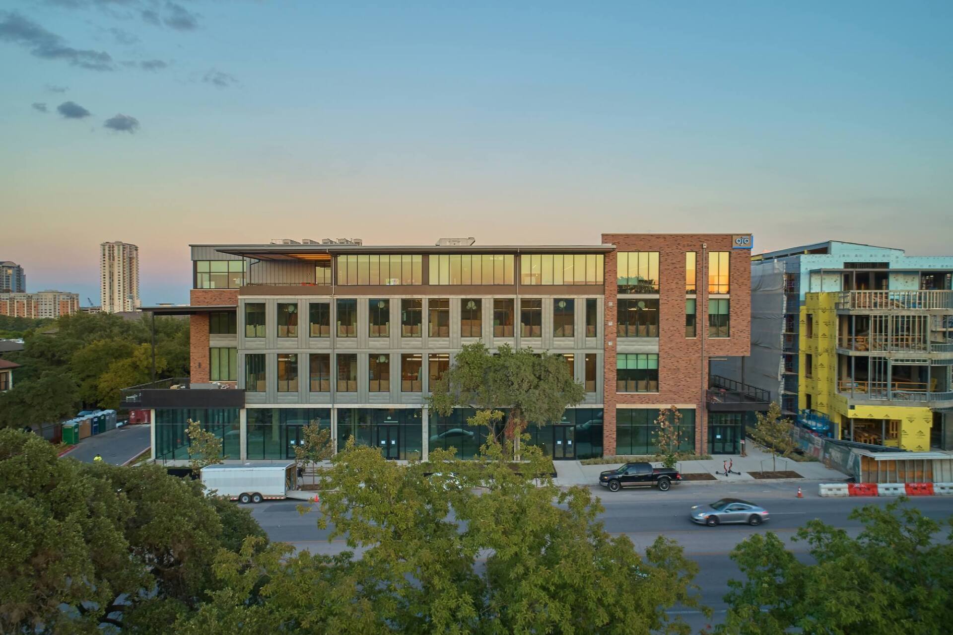 Multi-story brick and glass building with retail space on the ground floor, tree in the foreground, against a dusk sky.