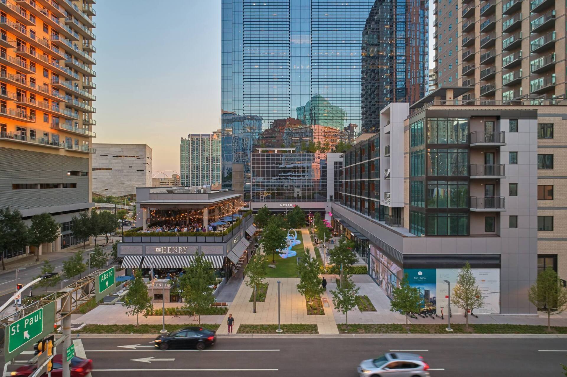 City street scene with buildings, restaurants, and a green space. Cars on the road.