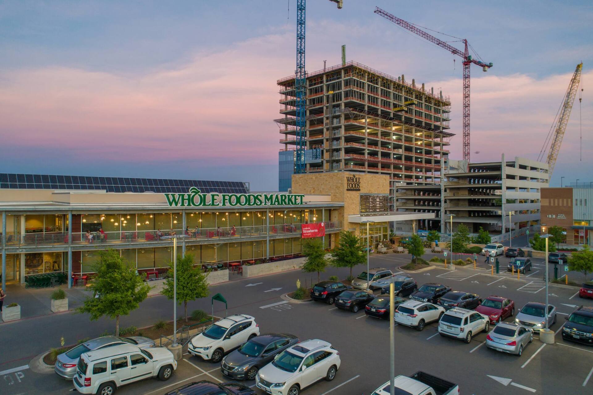 Whole Foods Market with a building under construction, parked cars, and cranes at dusk.