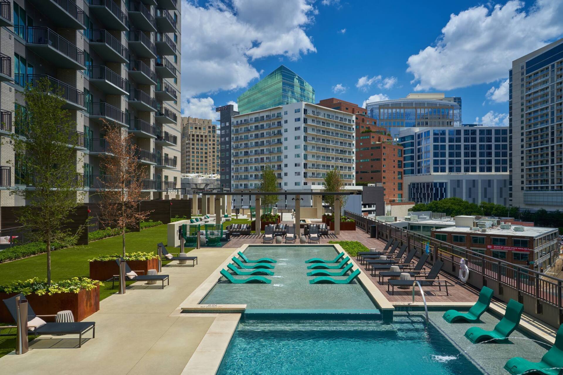 Rooftop pool and lounge area with city skyline backdrop, blue water, lounge chairs, green cushions, sunny day.