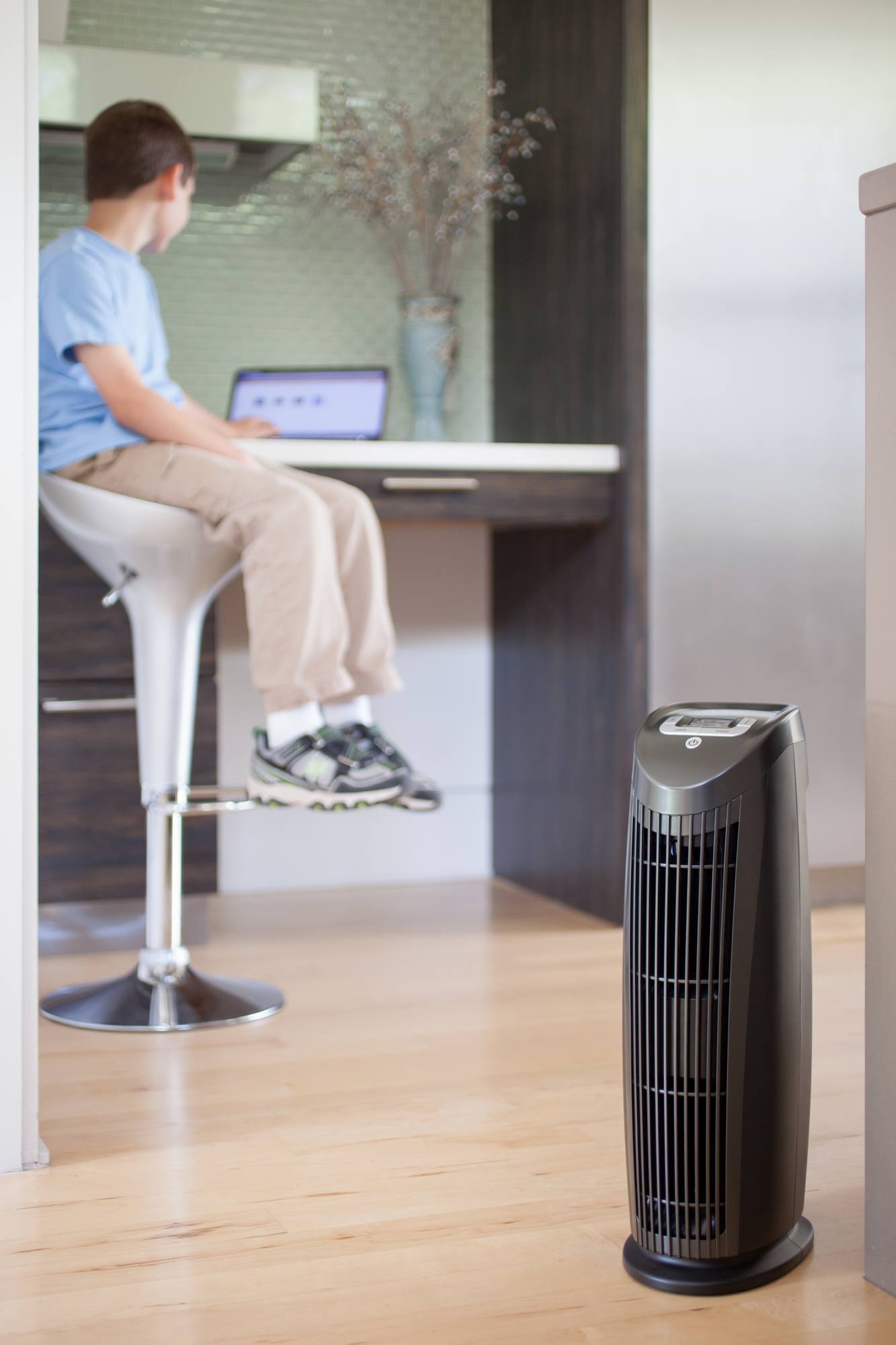 Black air purifier on hardwood floor; person working on laptop at counter.