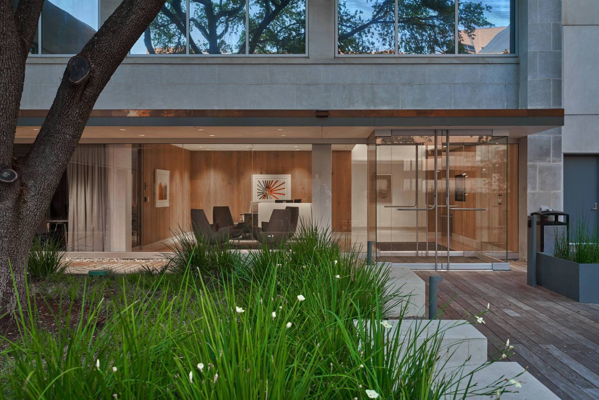 Exterior view of a modern building with glass doors, a seating area, and lush green landscaping.
