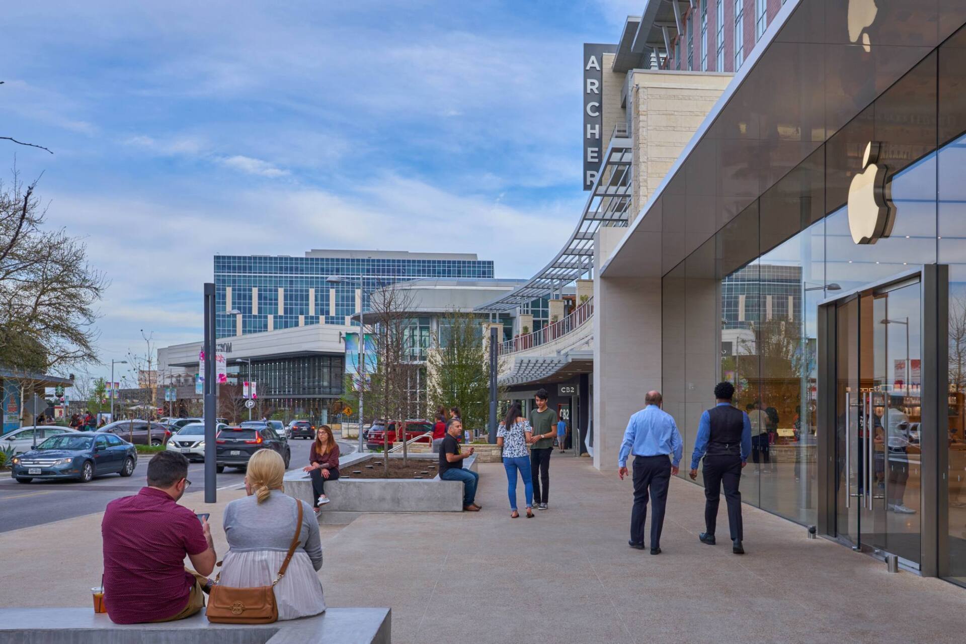 People walking outside an Apple Store with a large logo on the right. Others sit or walk on the sidewalk.