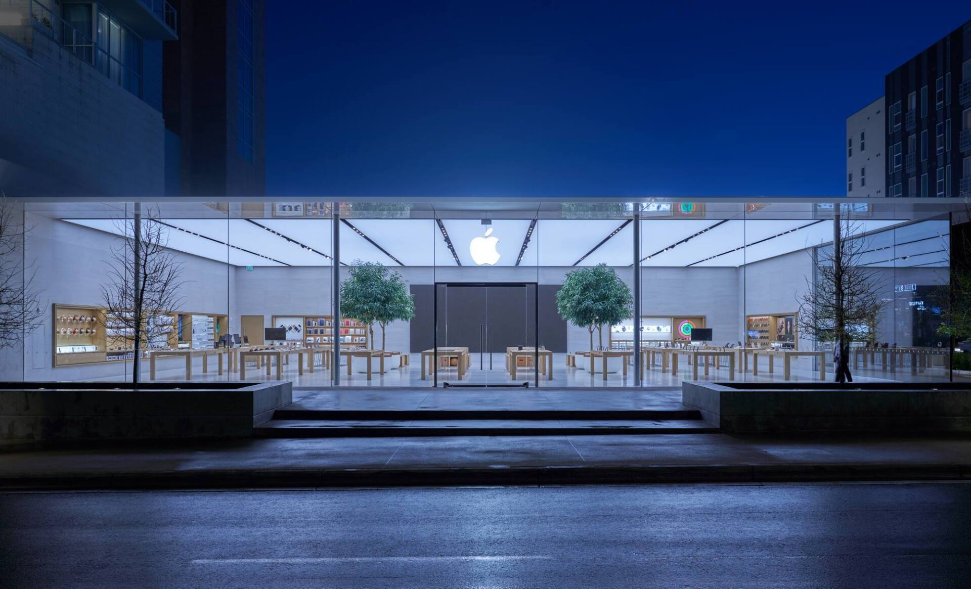 Apple store at night with glass facade, glowing interior, and trees outside.