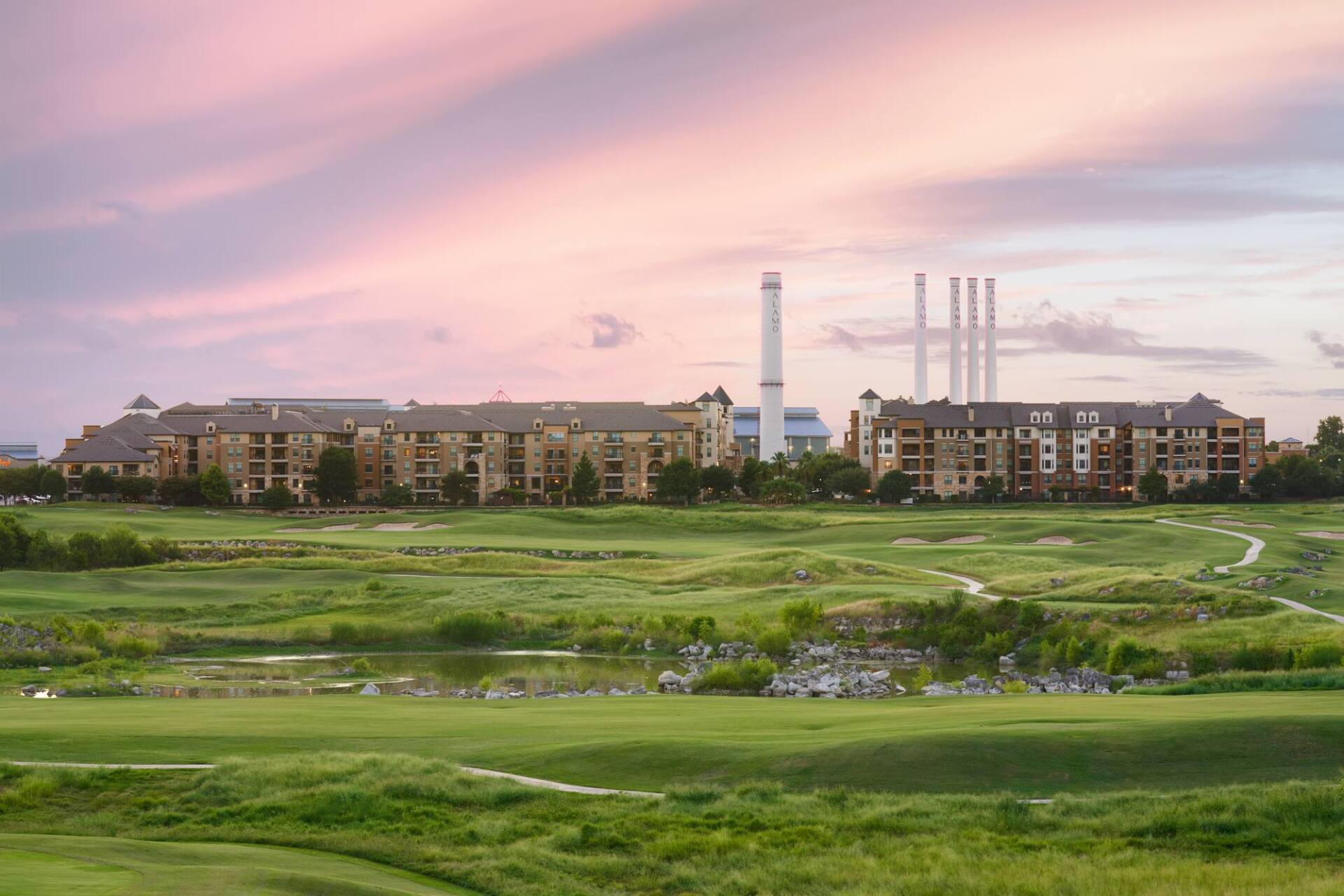 Golf course with green grass, buildings and power plant under a pink and purple sunset sky.