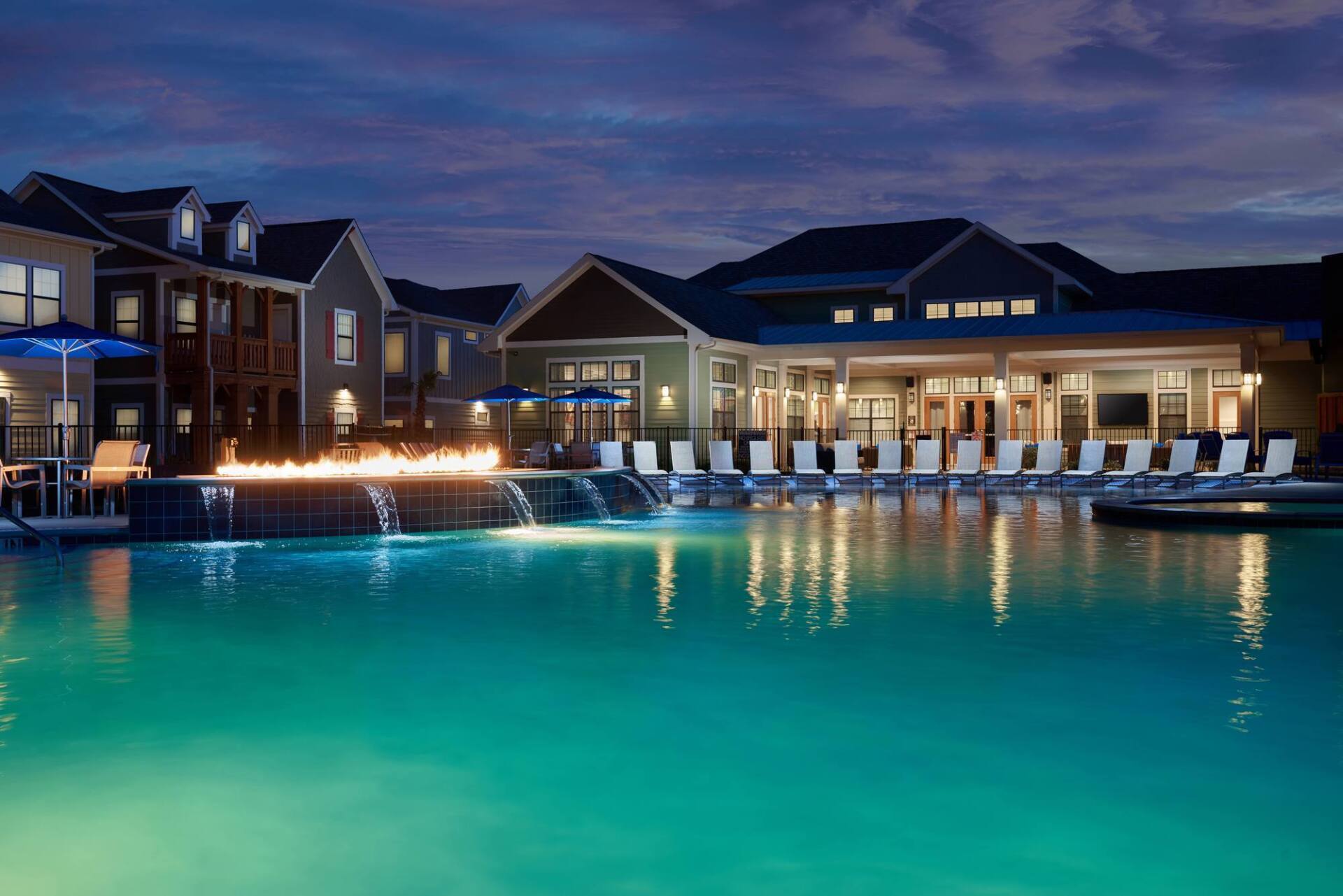 Night scene: Swimming pool with fire feature, lounge chairs, and buildings with lights.