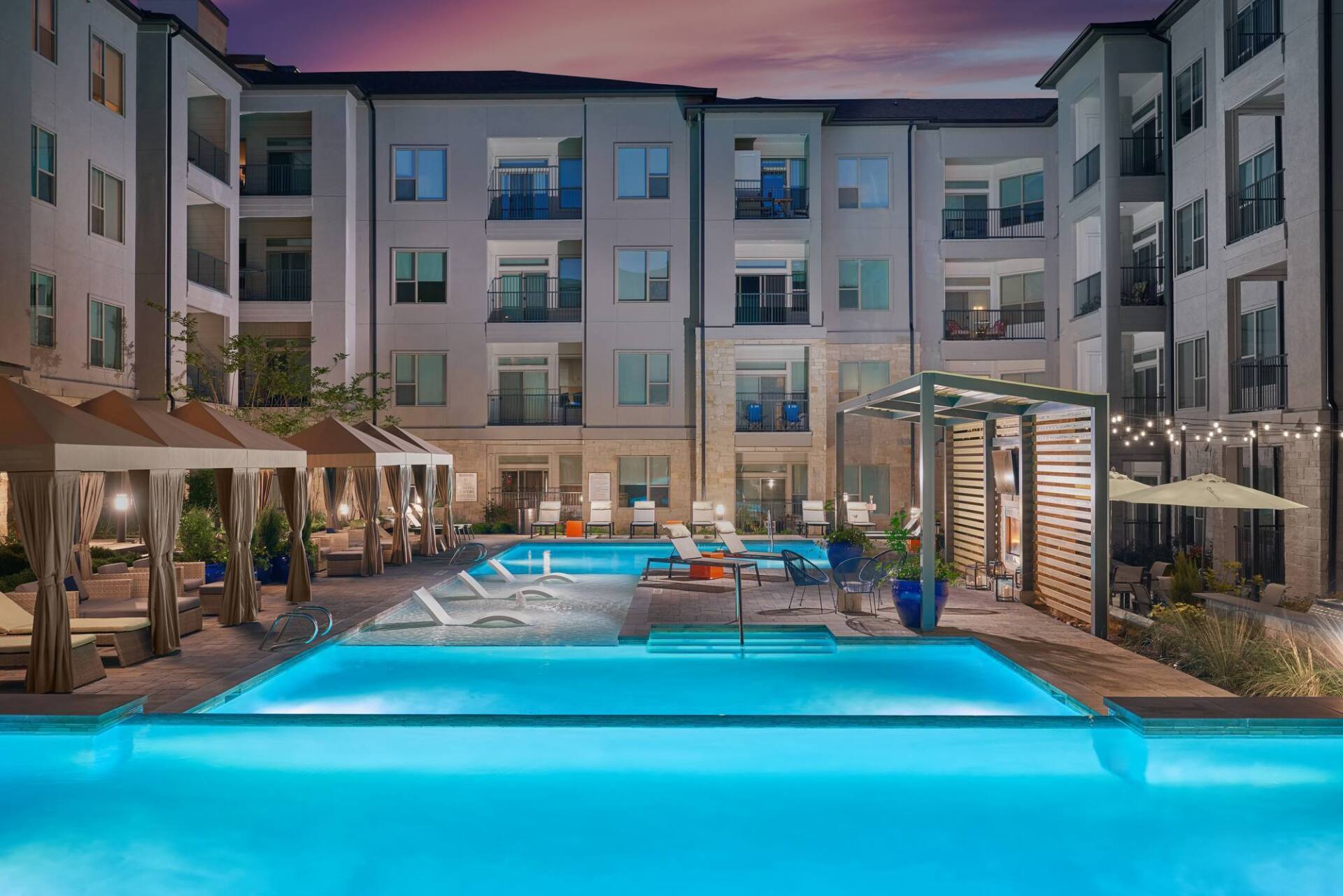 Apartment complex pool at dusk, with cabanas, lounge chairs, and building with balconies.