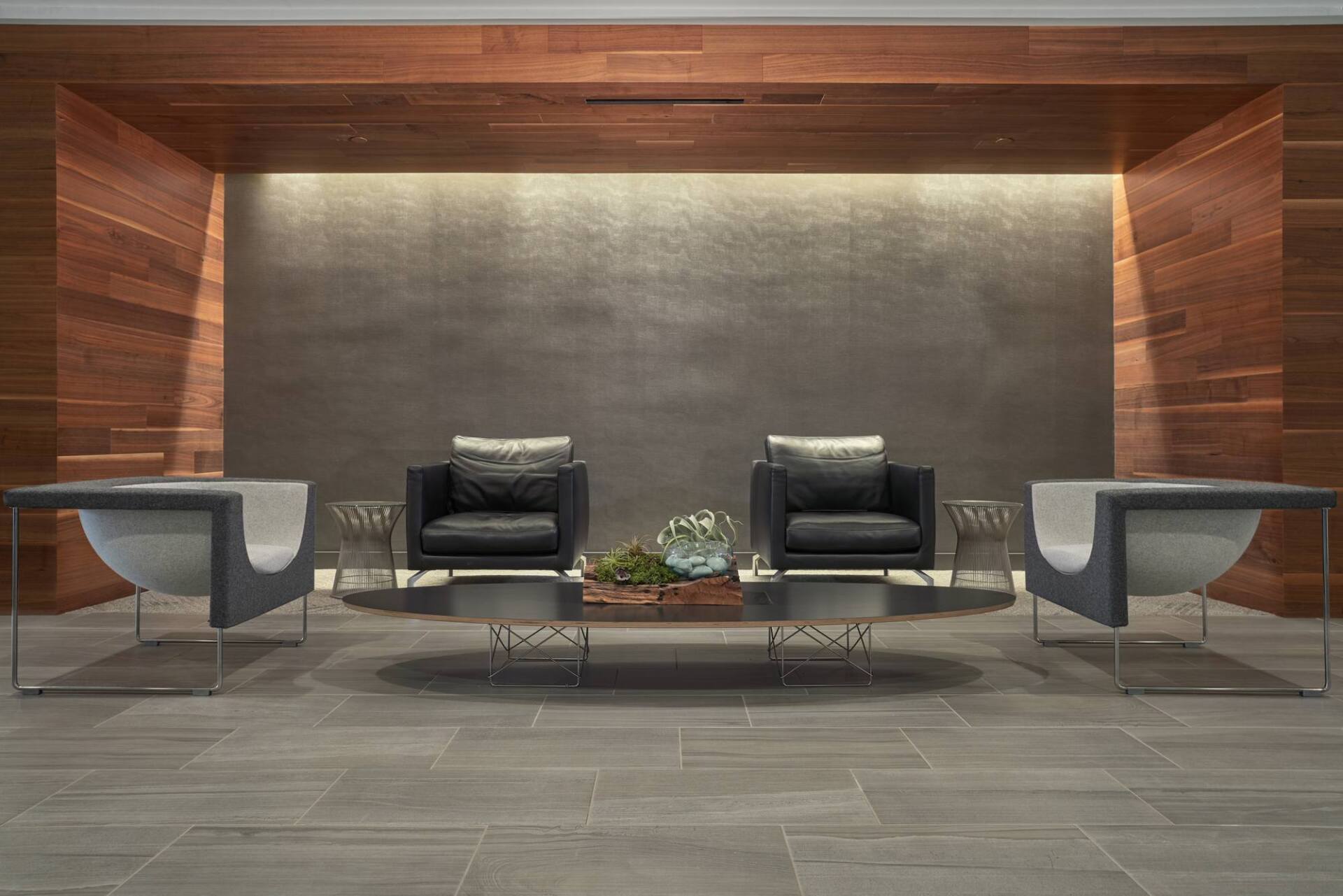 Modern lounge area with two black leather chairs, two curved grey sofas, and a glass coffee table.