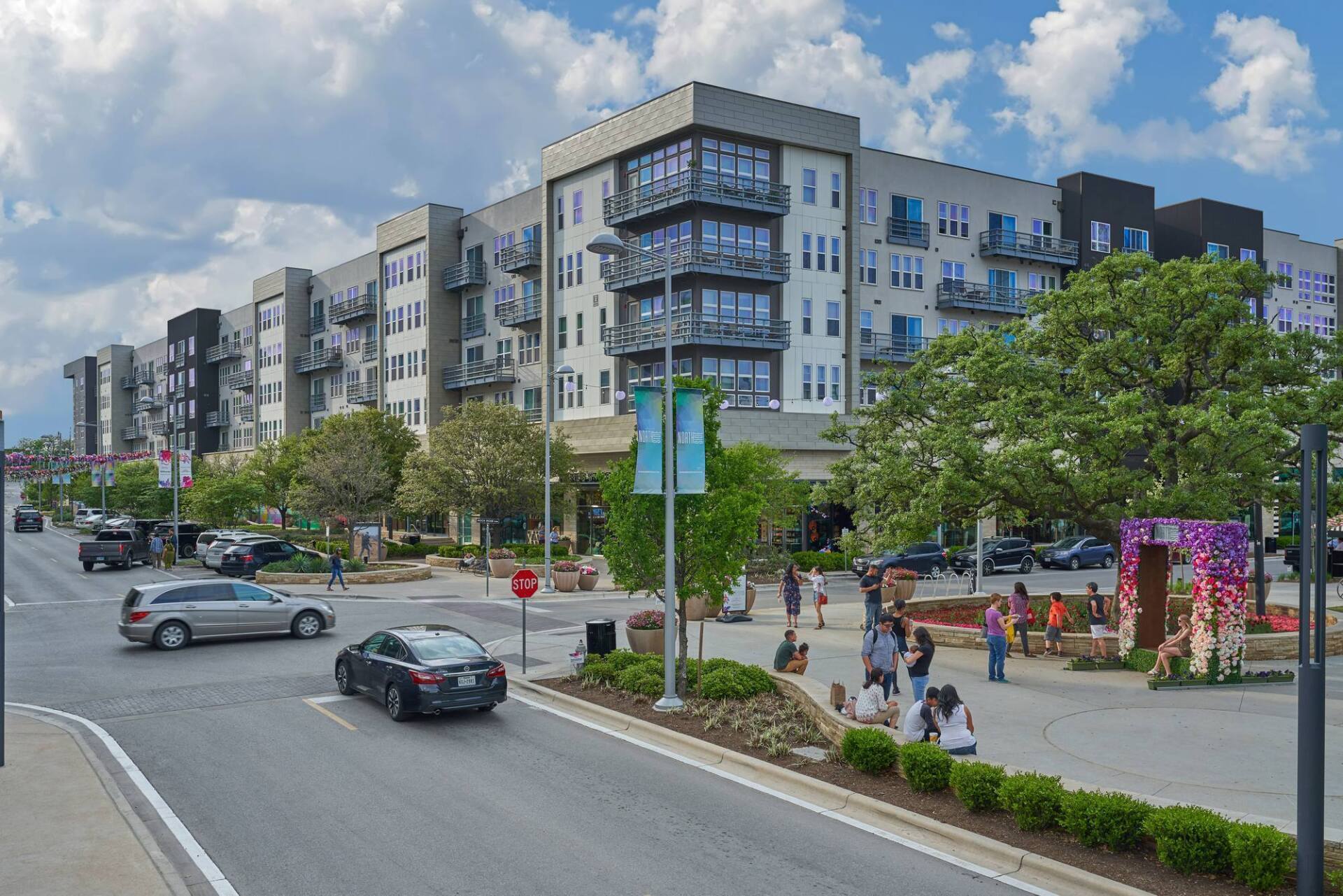 Multi-story apartment building with street, cars, pedestrians, and park area.