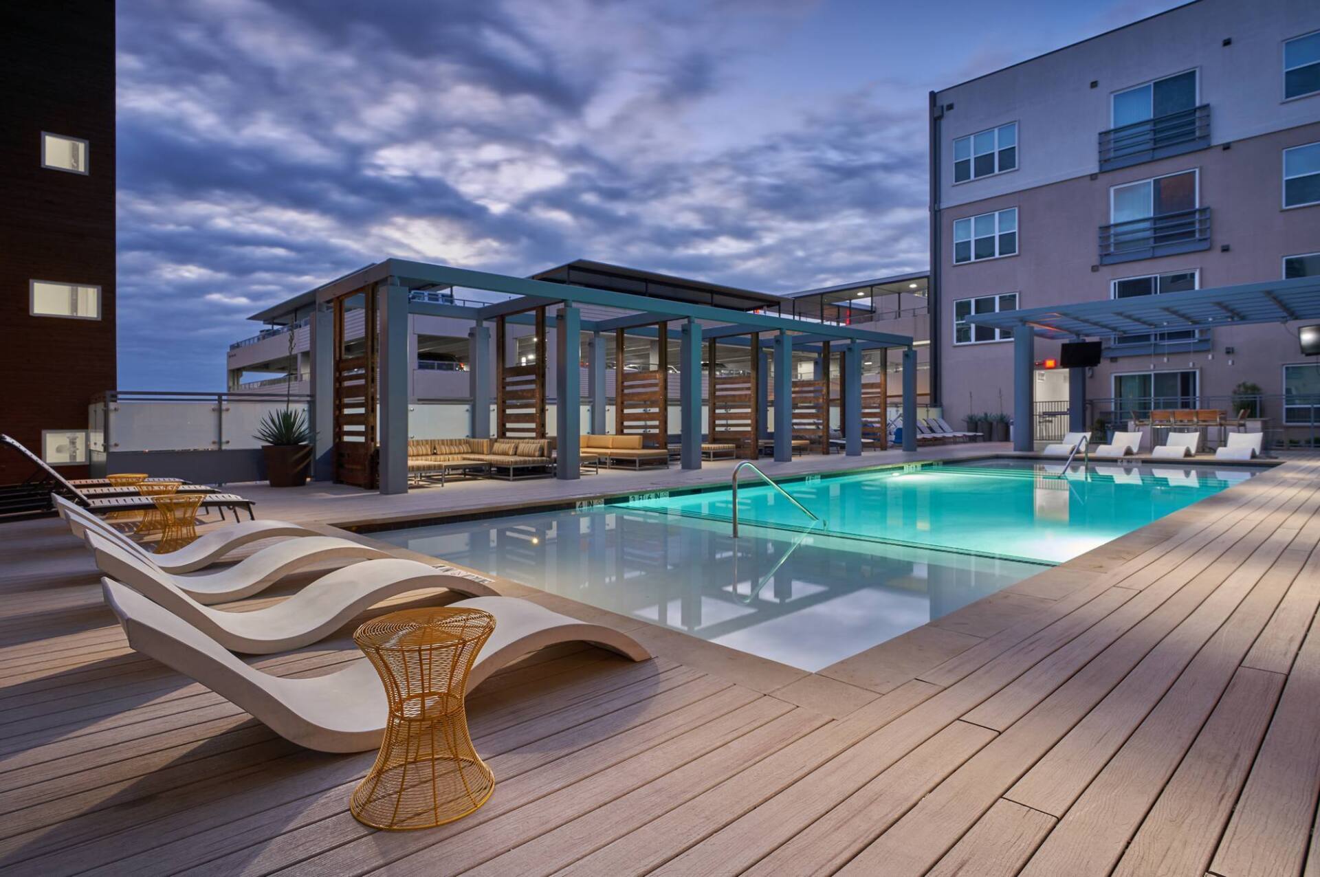 Pool with lounge chairs and pergola under a cloudy dusk sky. Apartment building in the background.