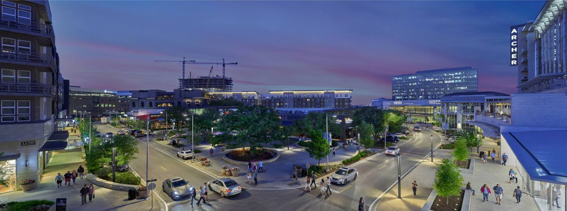 An urban scene at dusk. Buildings, streetlights, trees, and people. A construction crane is visible.