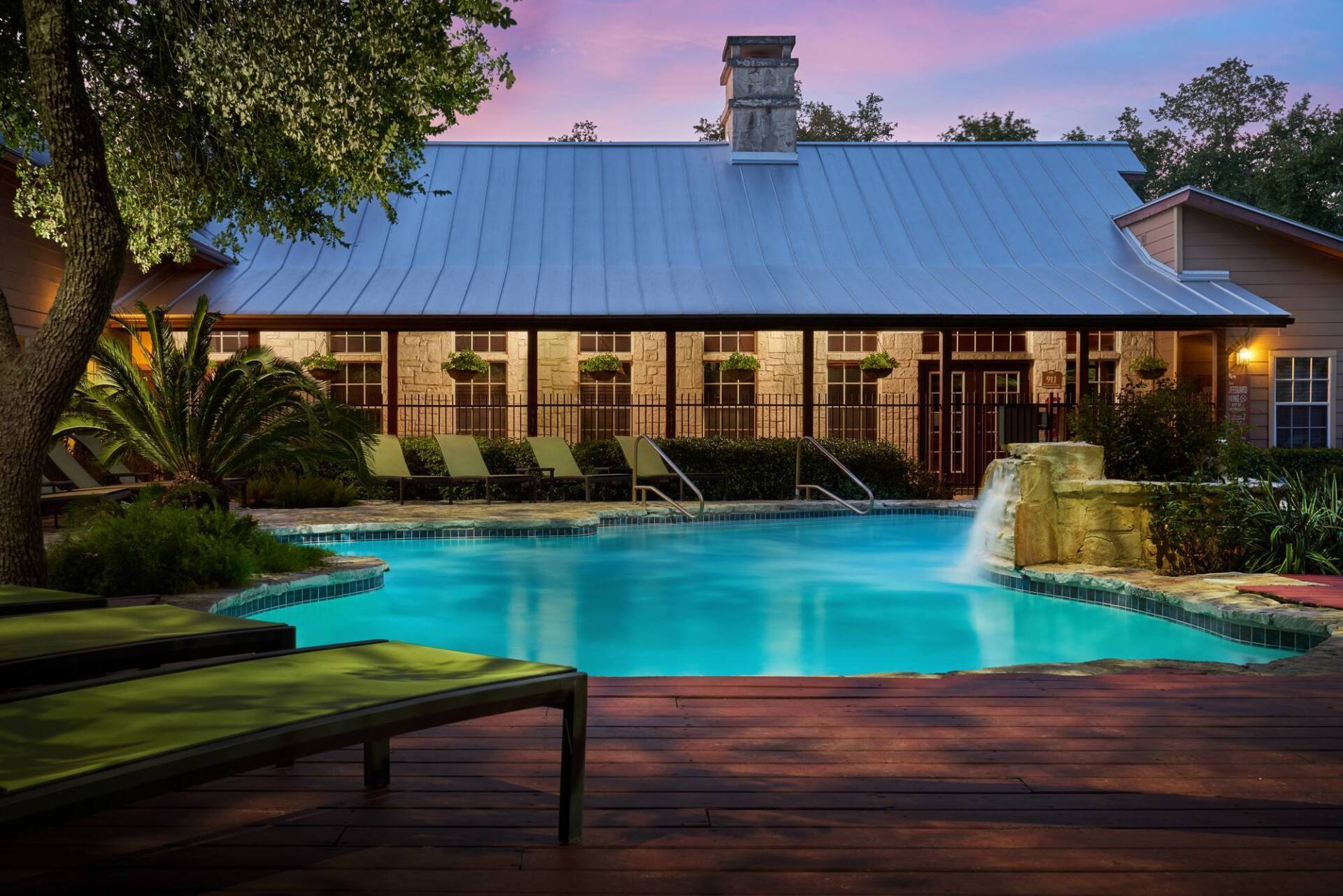 Swimming pool in front of a building with lounge chairs; waterfall feature, twilight sky.