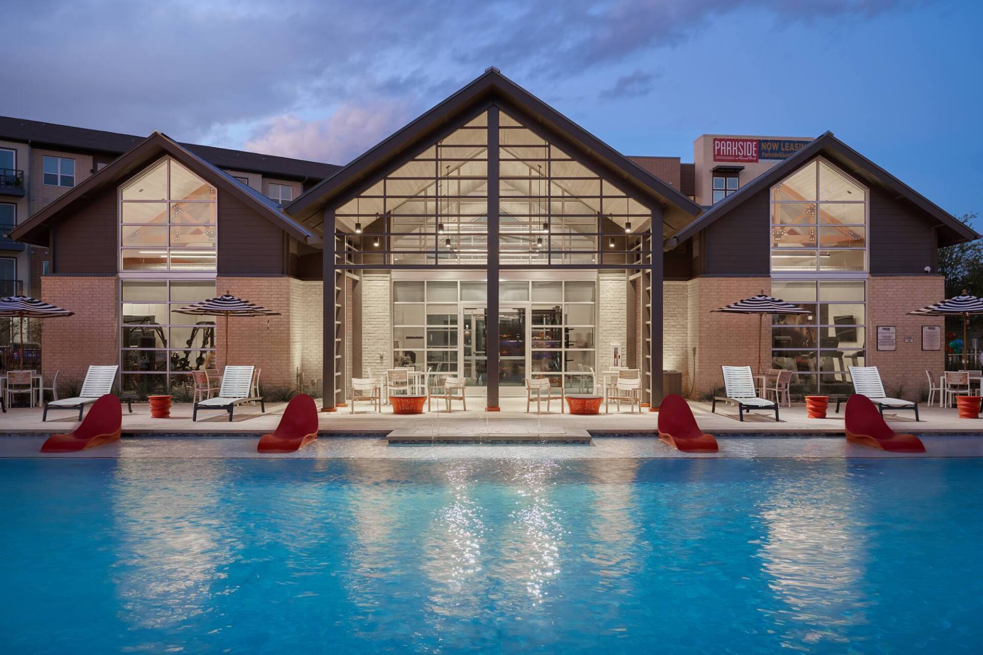 Pool in front of a modern building with glass doors and triangular roof, lounge chairs.