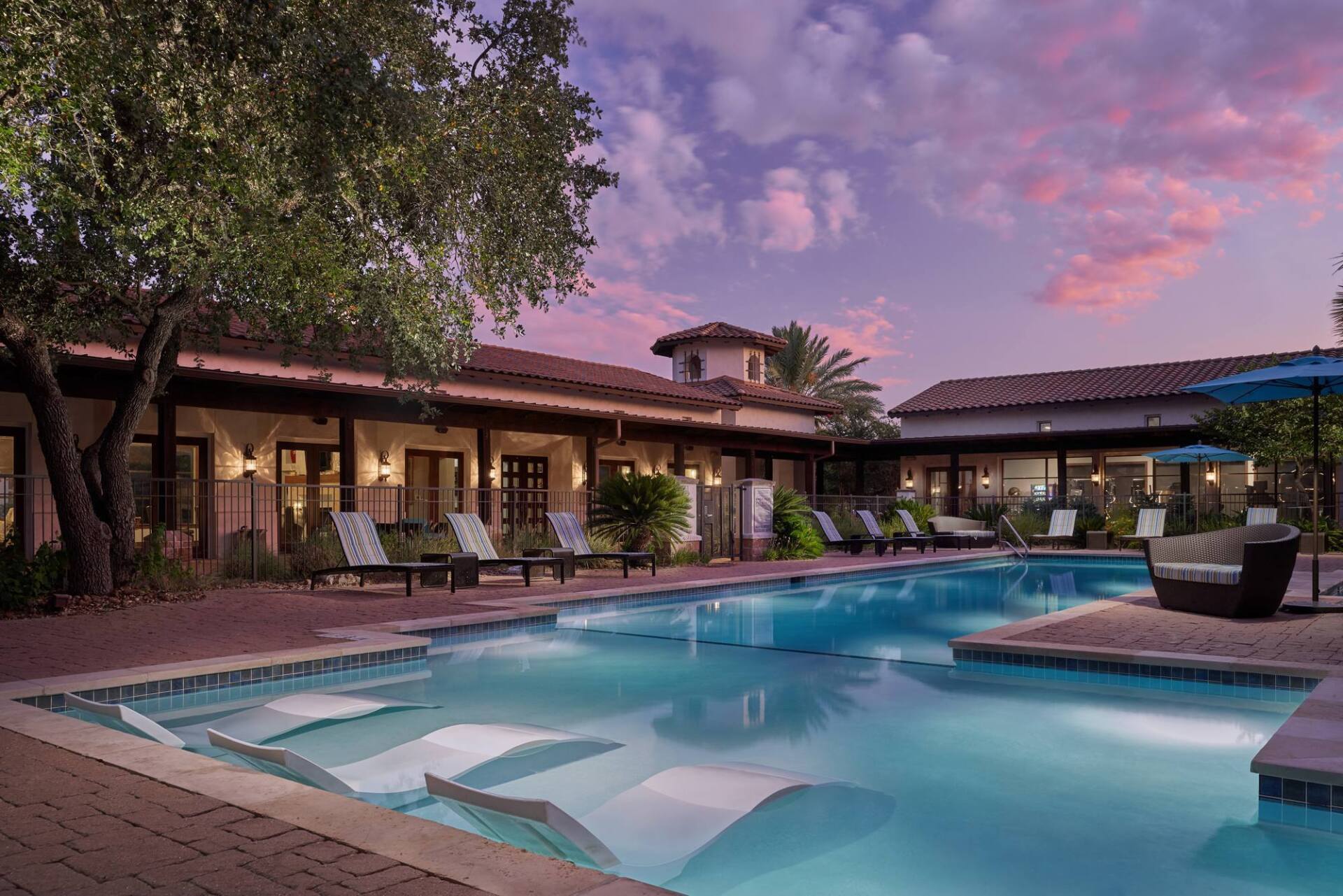 Pool at dusk with lounge chairs, building, and pink sky.