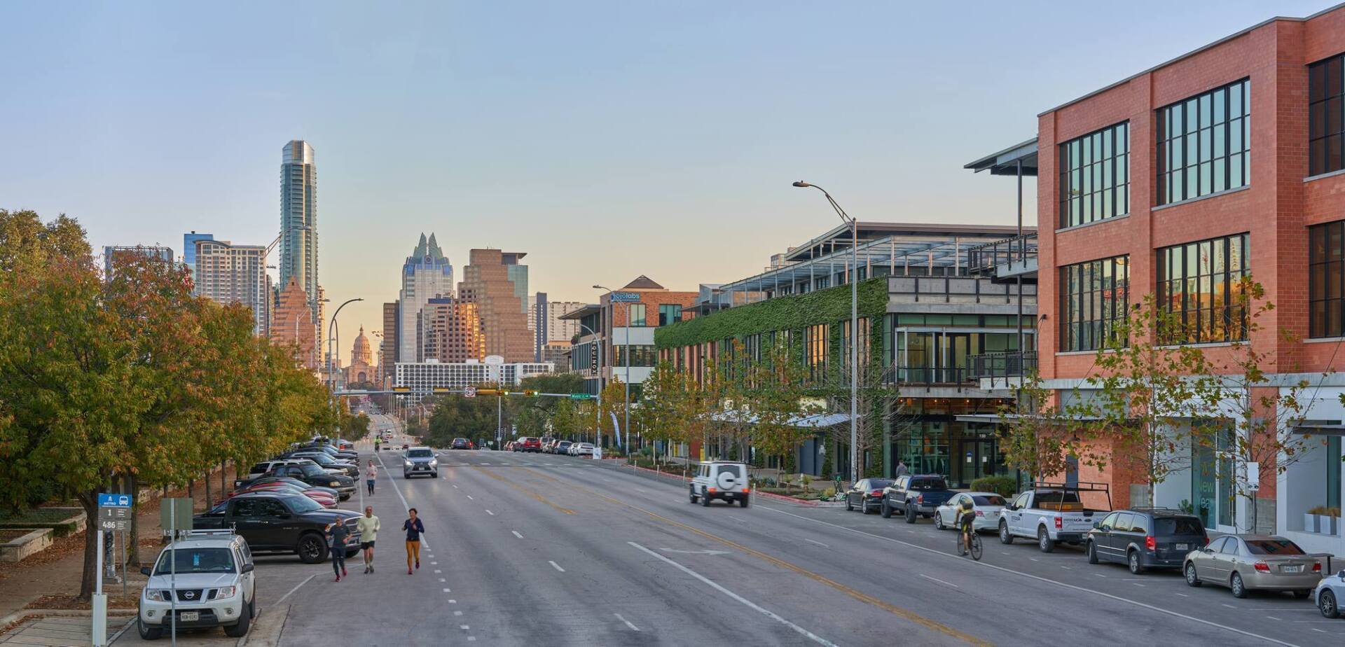 Street scene with buildings, vehicles, trees, and pedestrians. City skyline in background.