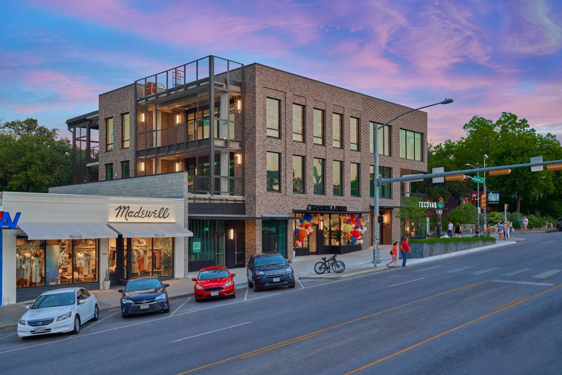 Multi-story brick building with street-level retail, cars parked out front, and a blue-pink sunset sky.