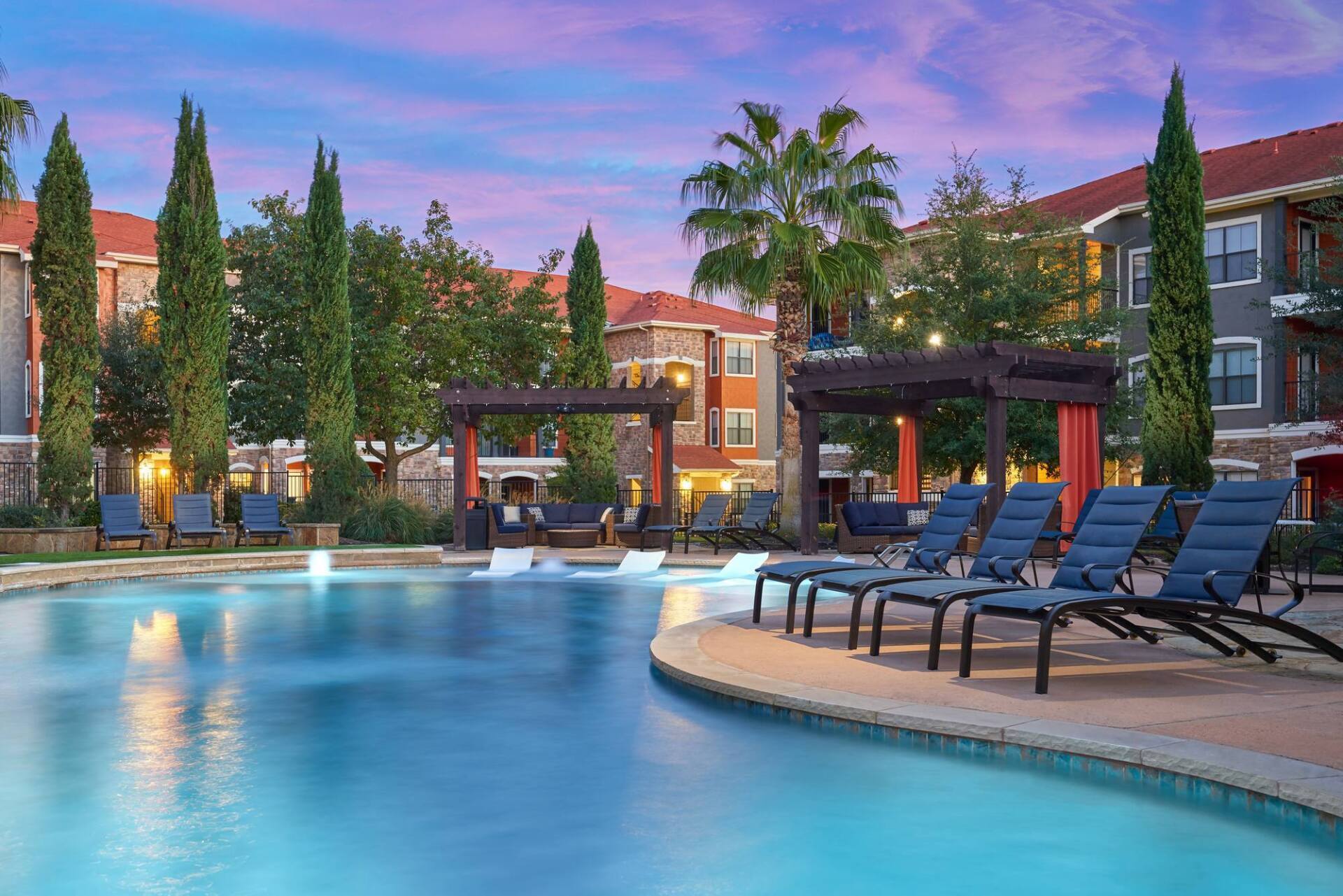 Pool with lounge chairs at an apartment complex under a twilight sky, surrounded by trees and buildings.