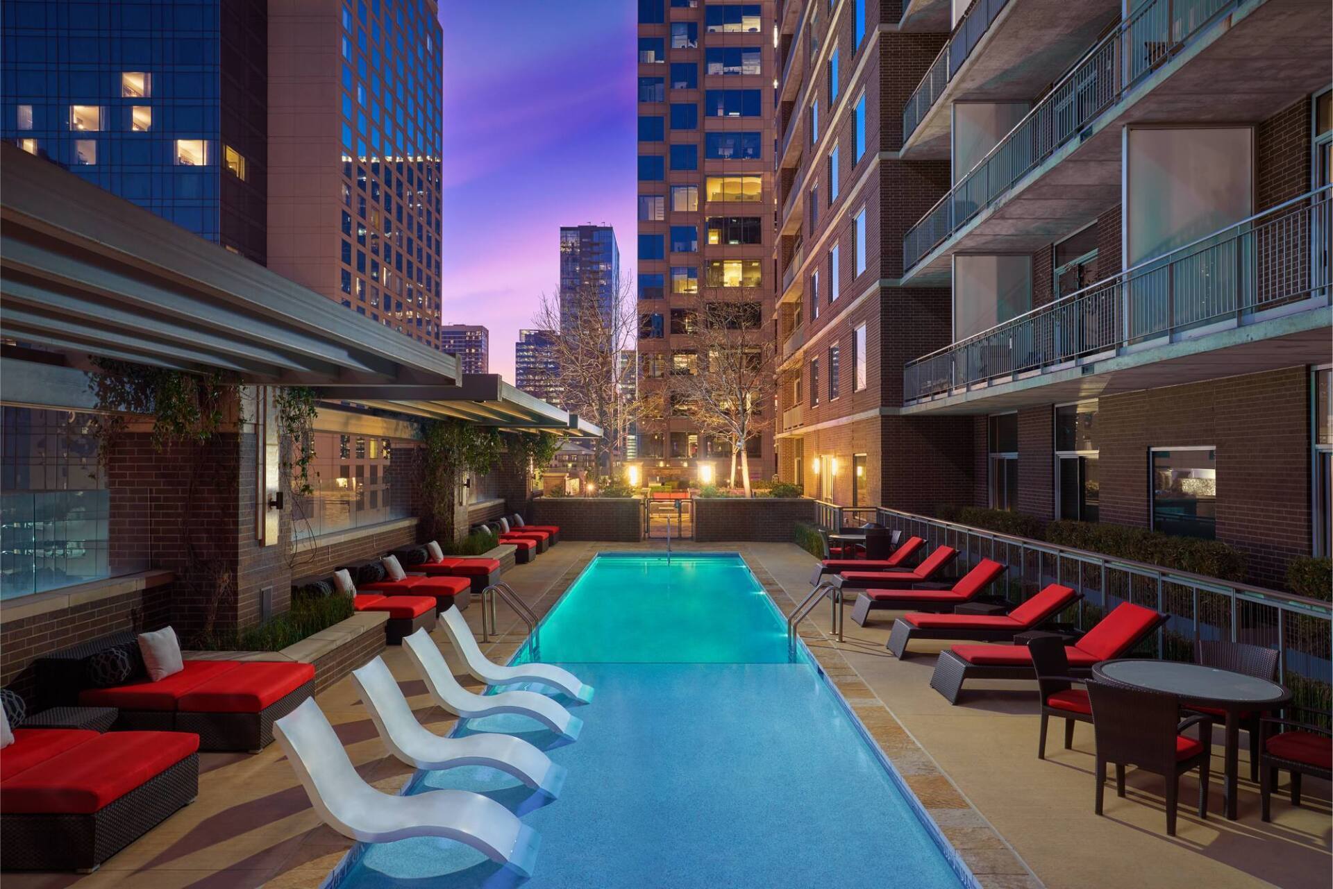 Poolside lounge area at dusk, featuring a rectangular pool, red lounge chairs, and high-rise buildings.