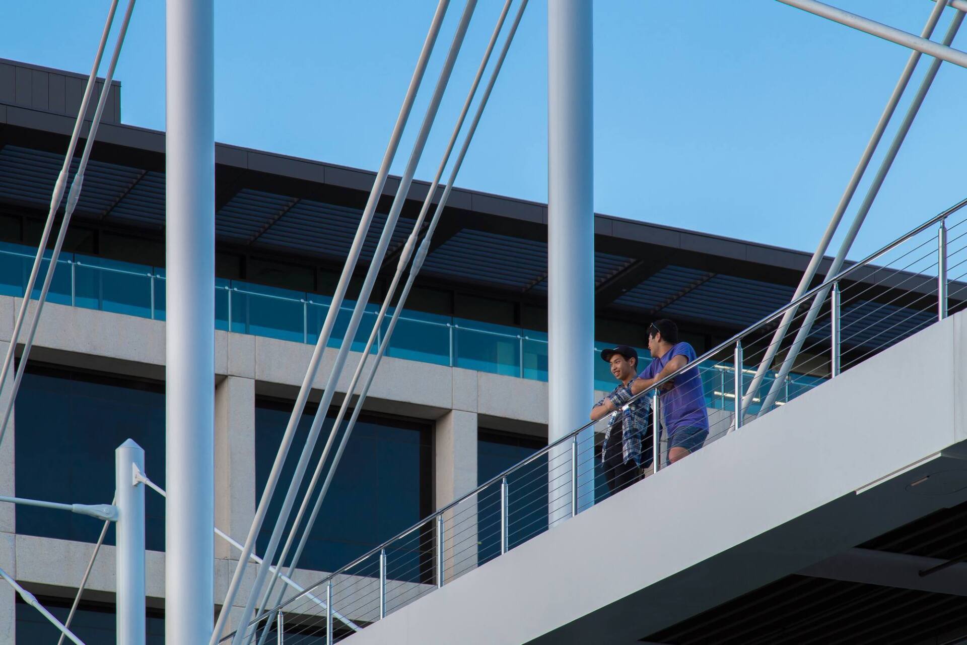 Two people leaning on a railing of a bridge-like structure with a building and sky in the background.